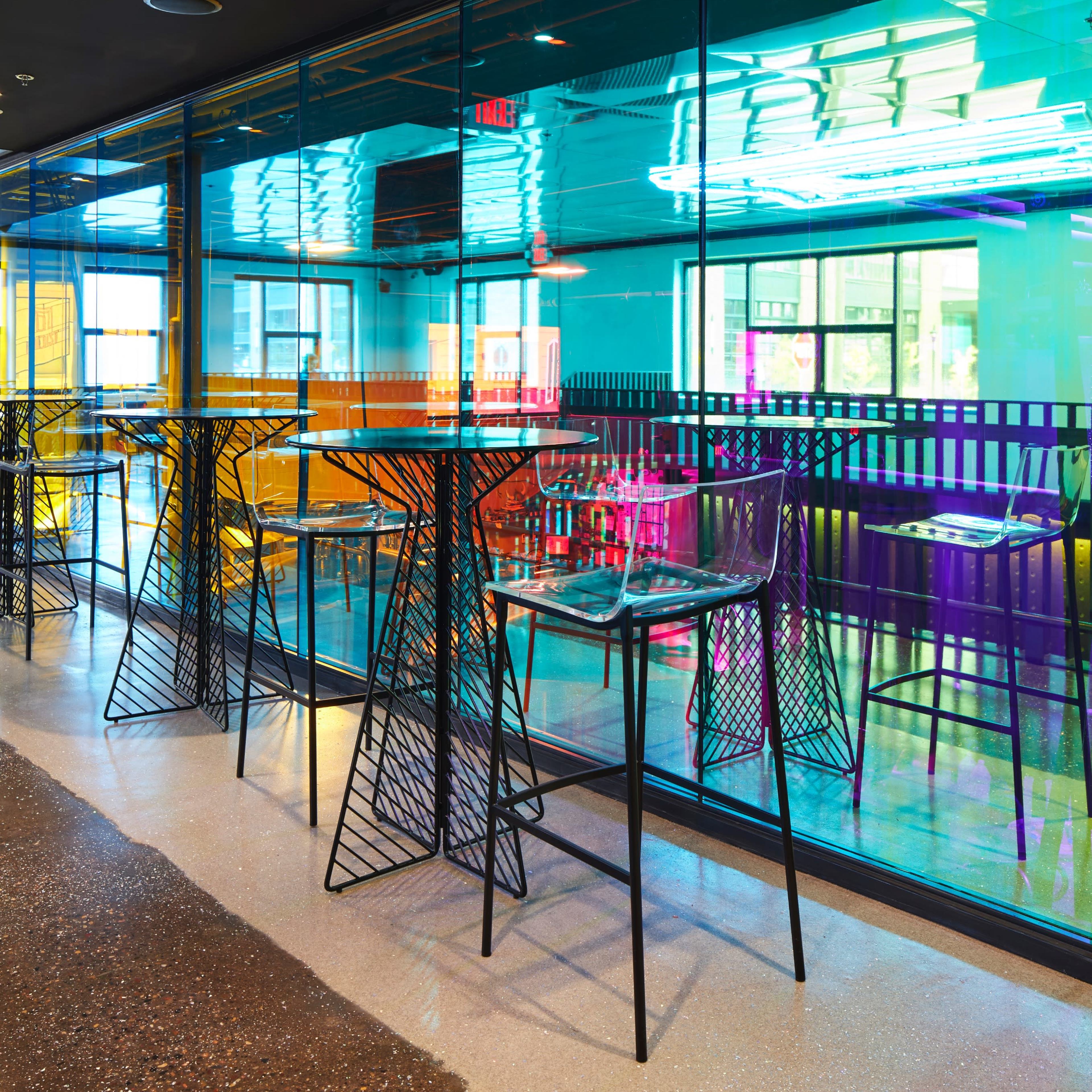 A modern cafe with colorful illuminated walls and transparent glass panels, featuring high black tables and chairs arranged in a spacious layout.