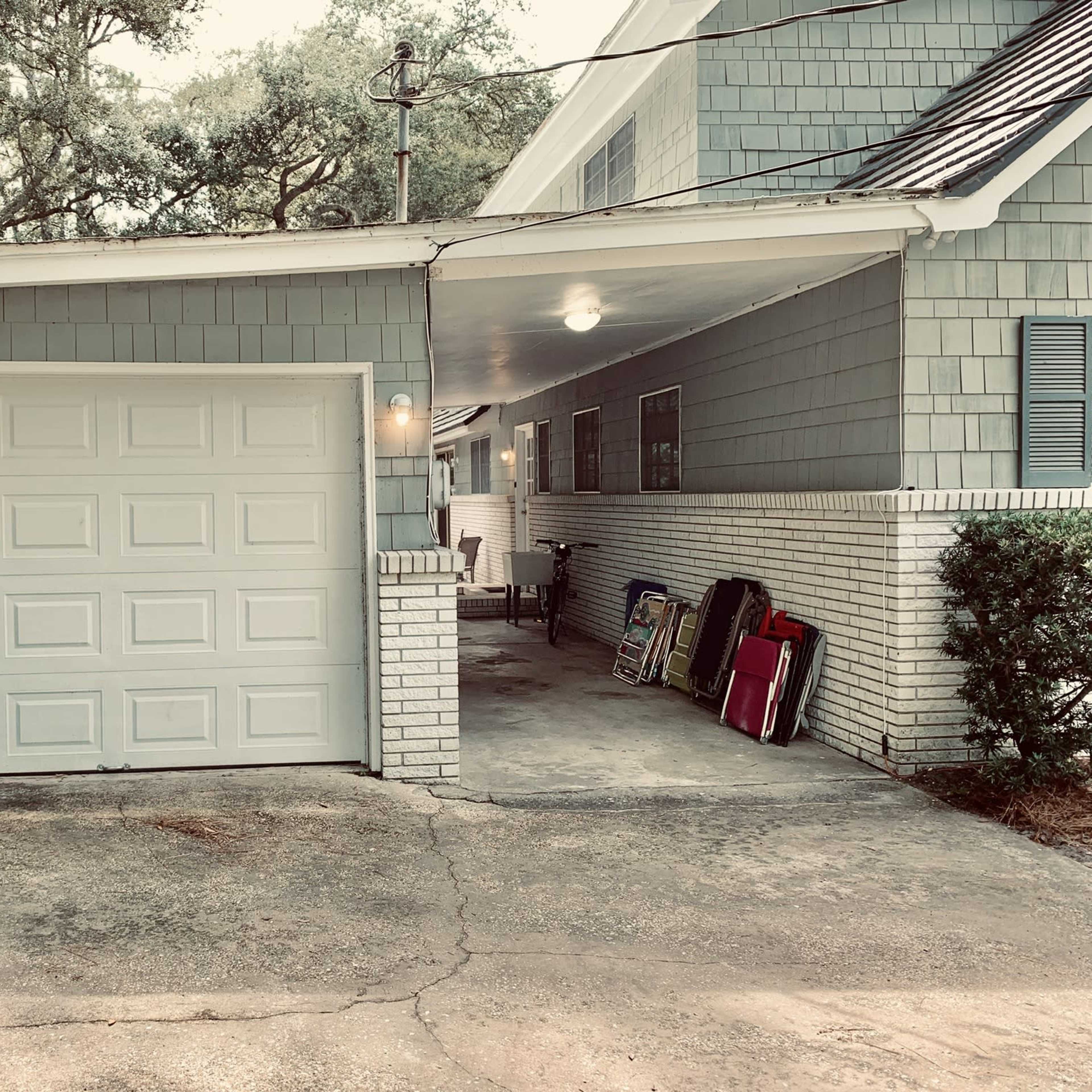 A garage with a white door, next to a green trash bin and a pathway leading to a storage area with stacked chairs.