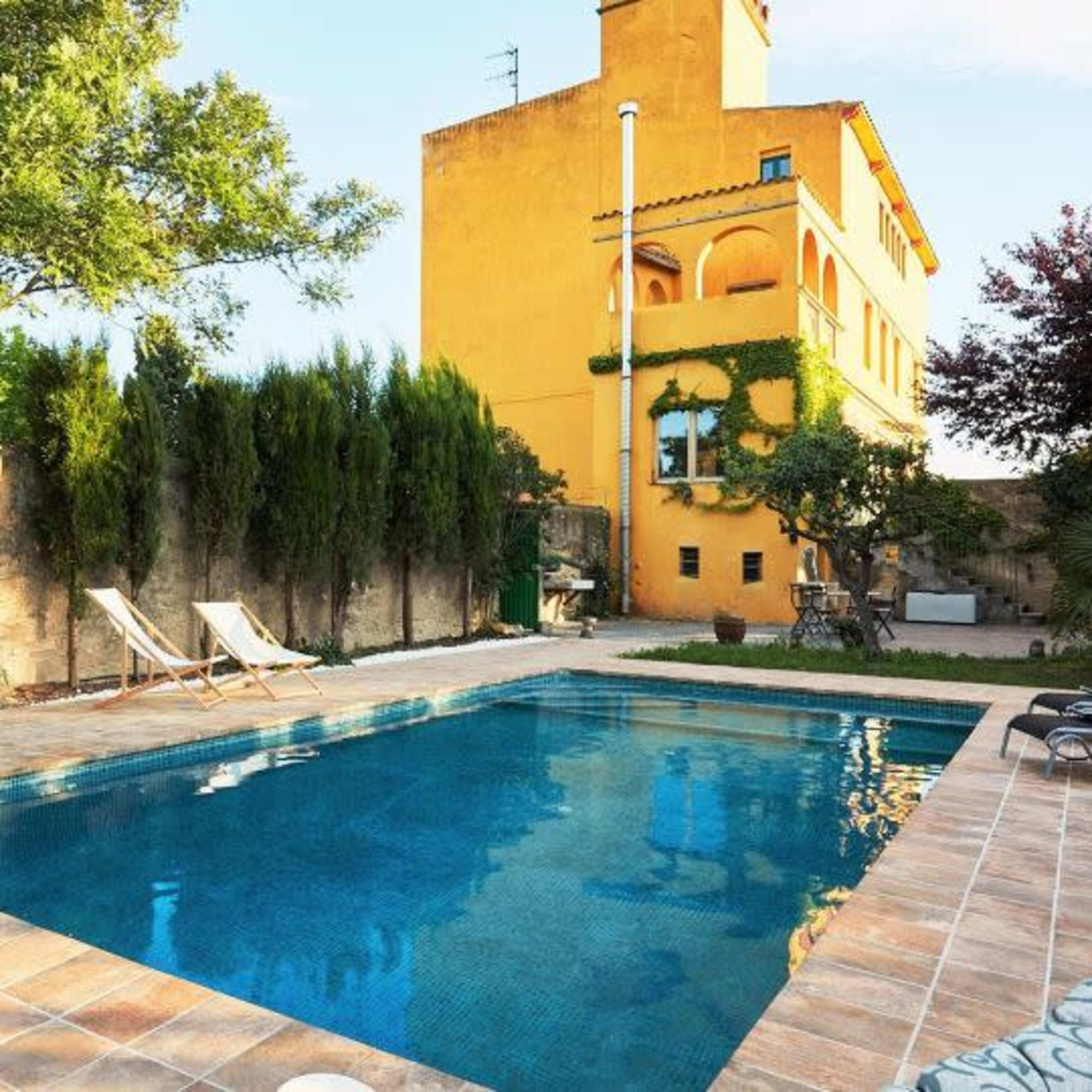 A rectangular swimming pool is surrounded by lounge chairs and greenery, with a yellow building in the background under a clear sky.