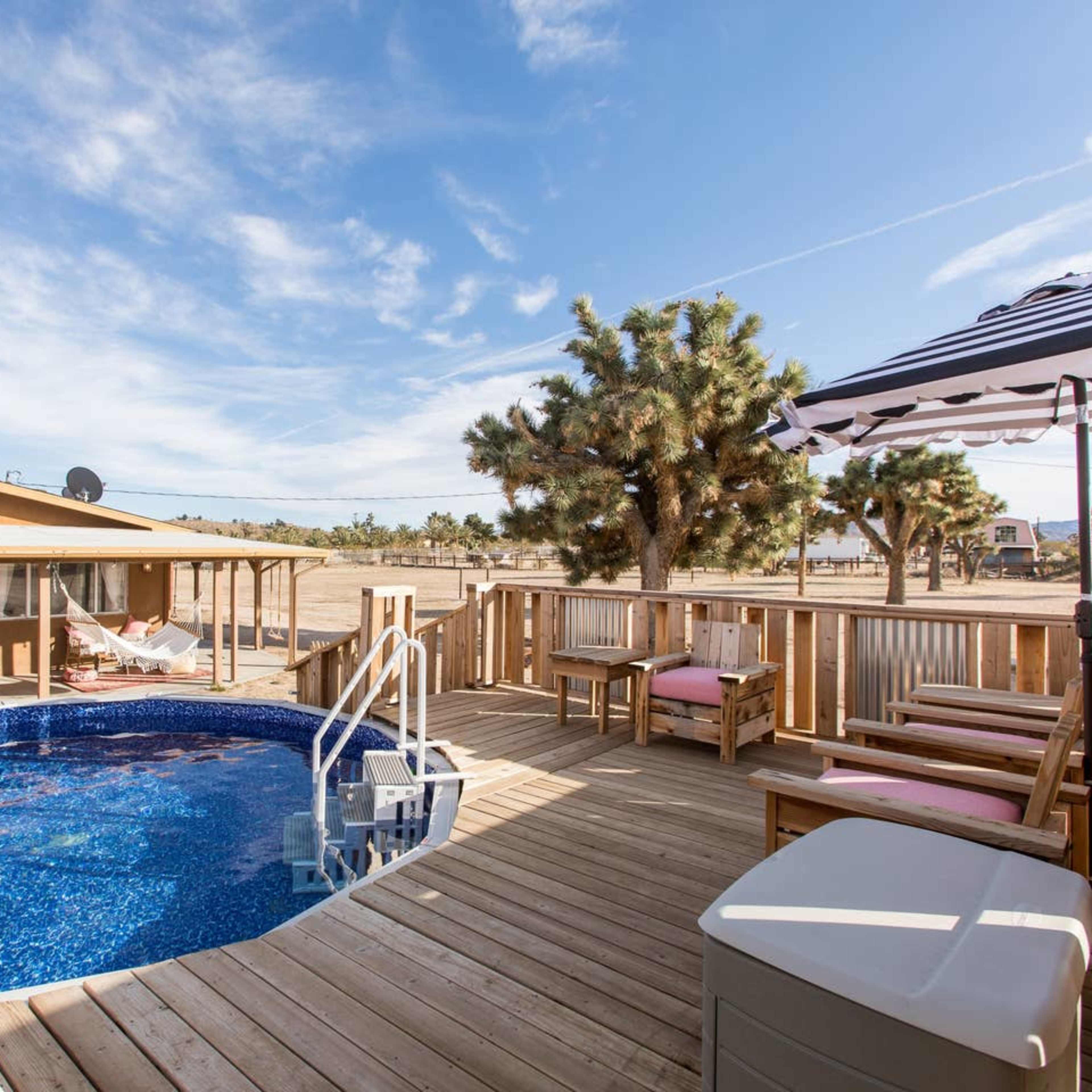 A wooden deck surrounds a round swimming pool adjacent to a single-story house, with seating areas and a shaded umbrella nearby.