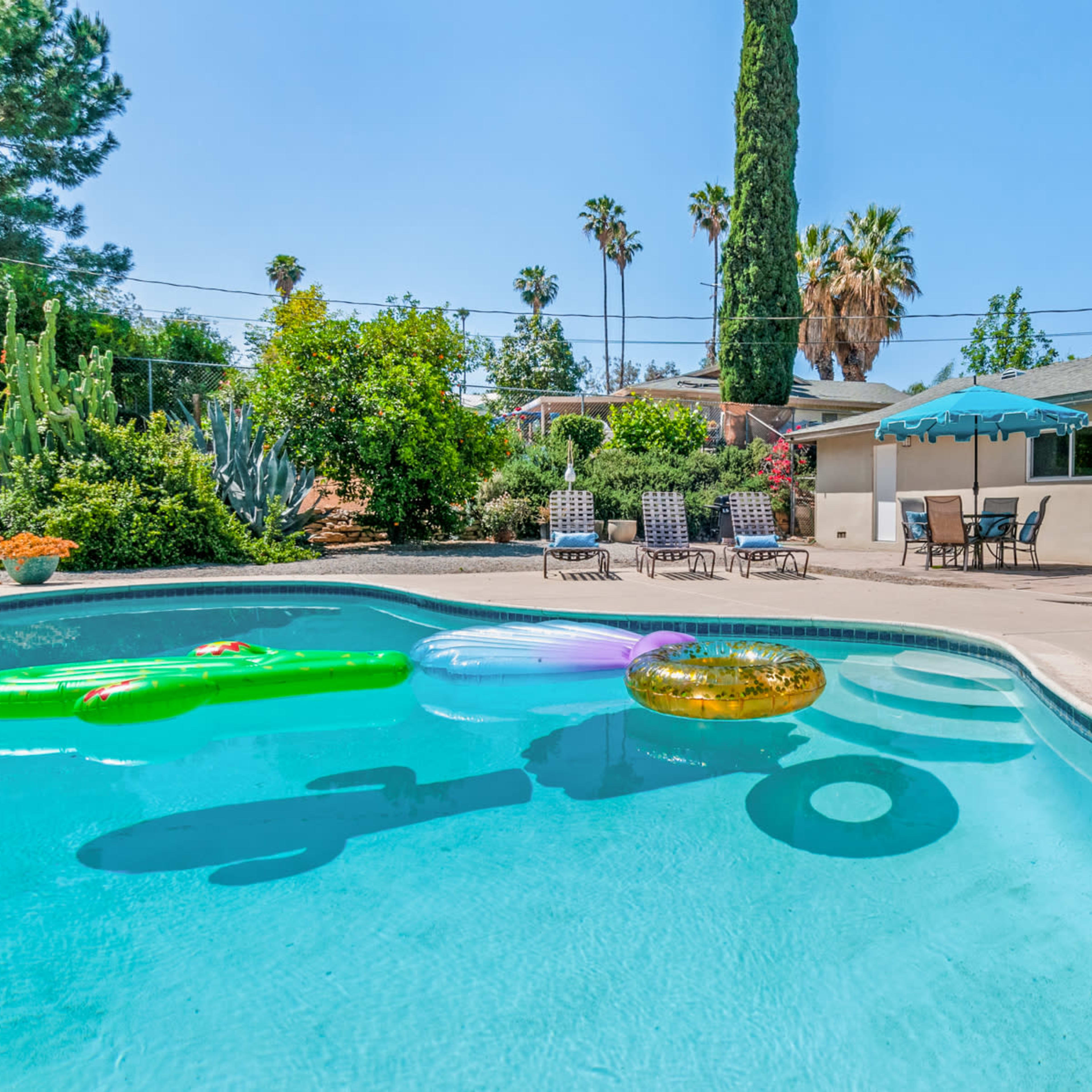 A swimming pool surrounded by lounge chairs and several colorful pool floats, with trees and shrubs in the background.