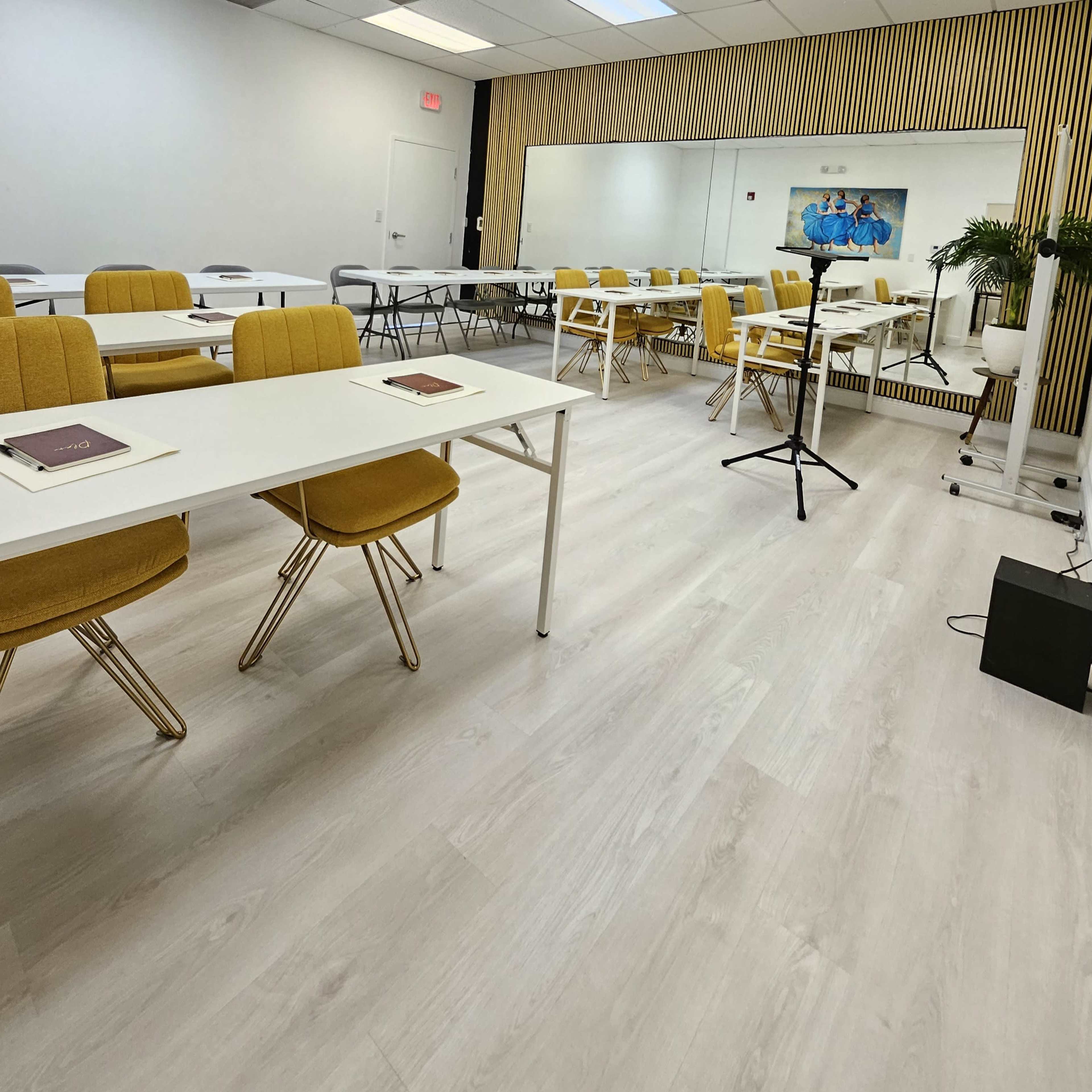 A bright, modern classroom setup with yellow chairs arranged around white tables and a large mirror on one wall.