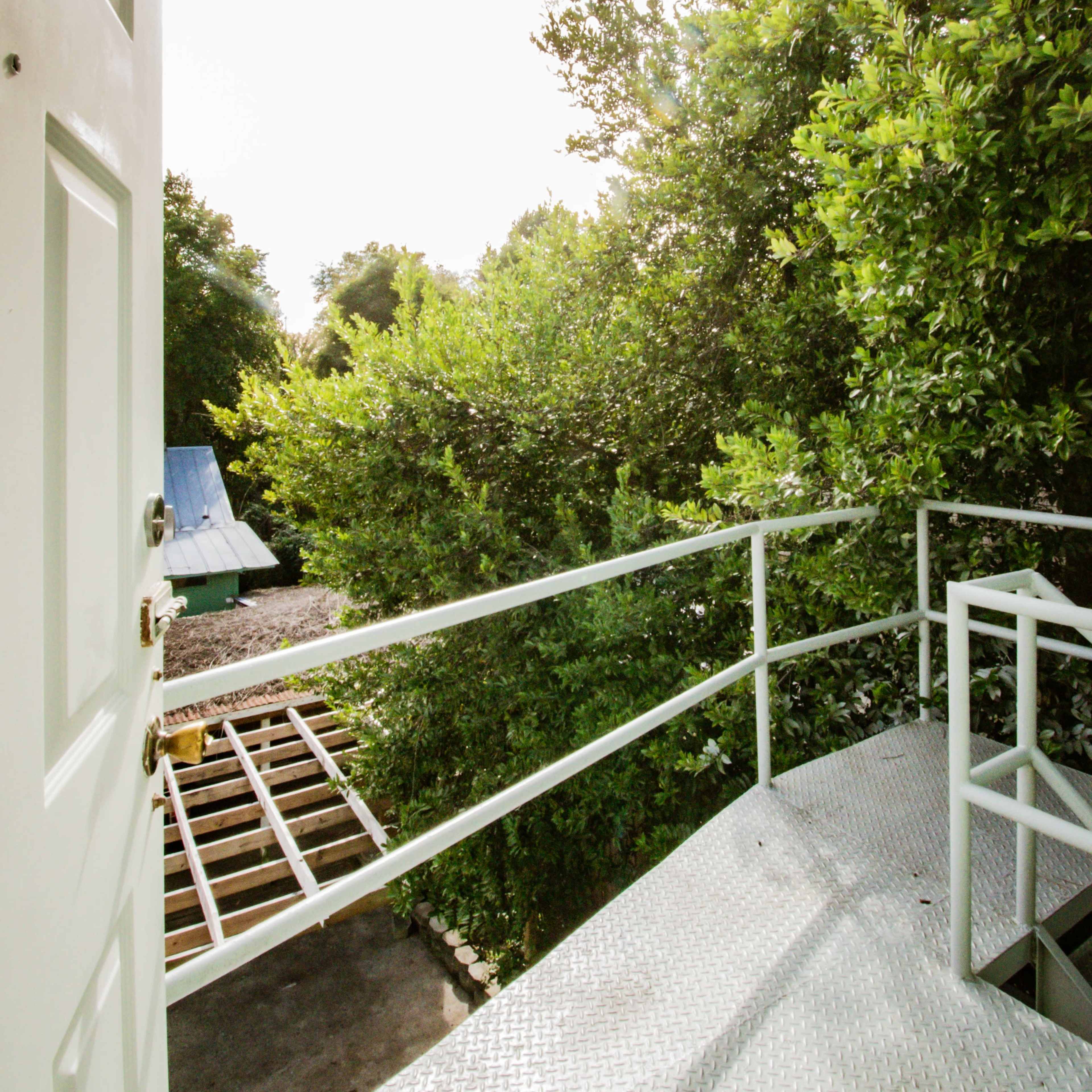 A metal door opens onto a narrow balcony surrounded by dense greenery, leading to a view of a wooden pathway and a partially visible structure in the background.