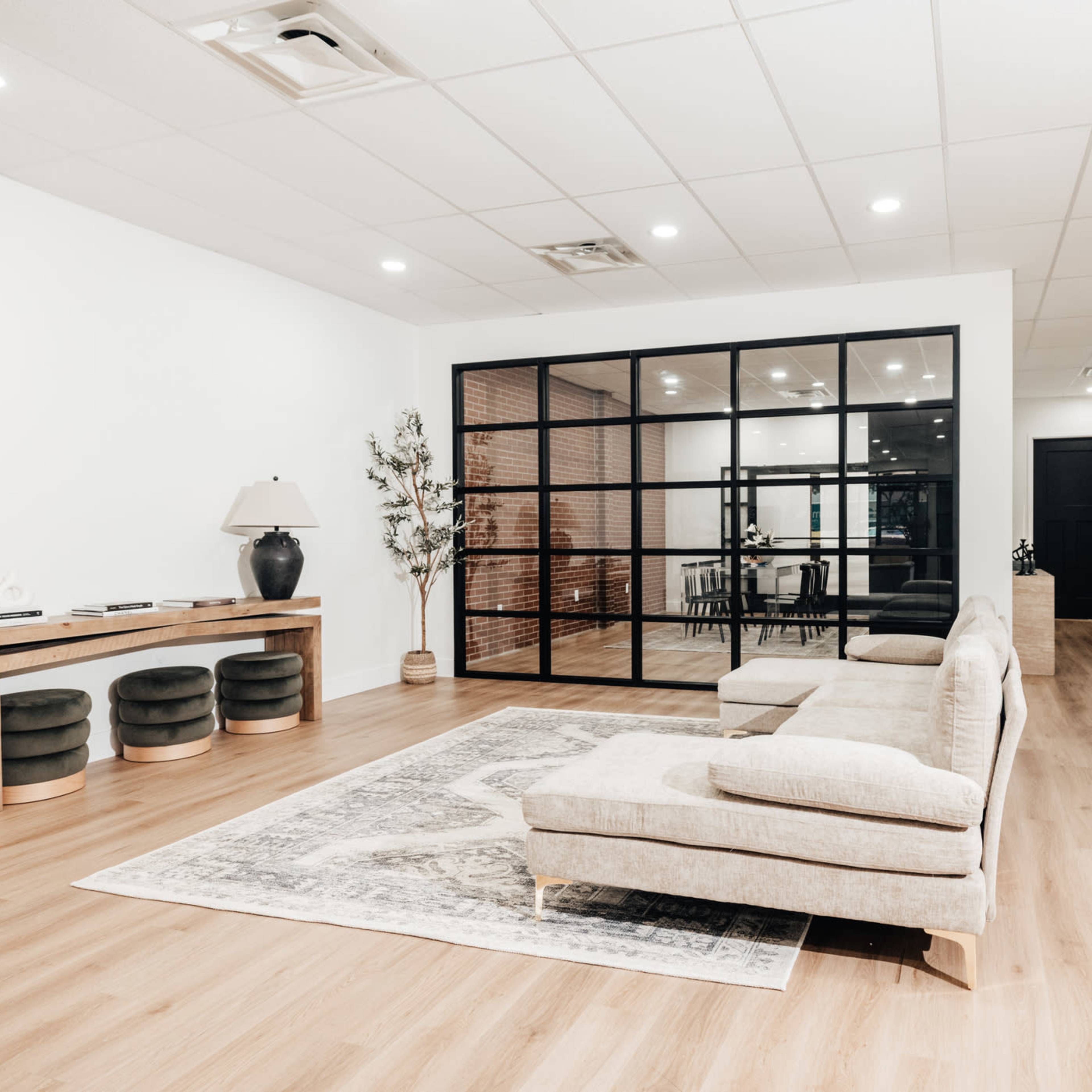A spacious room features a modern beige sectional sofa, a wooden console table, and a glass-enclosed meeting space in the background.