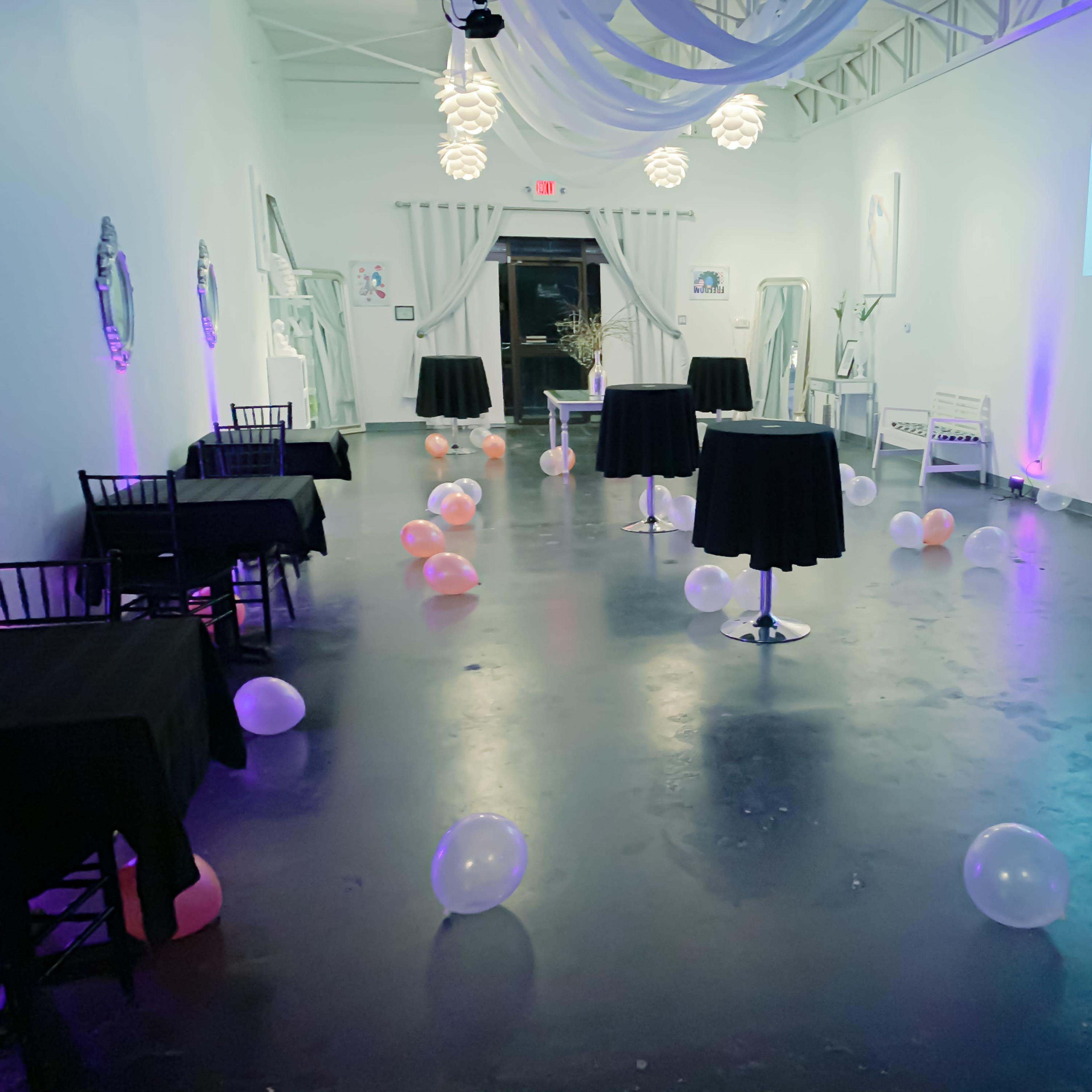 A spacious event room features several black-covered tables surrounded by scattered pink and white balloons under soft, colorful lighting.