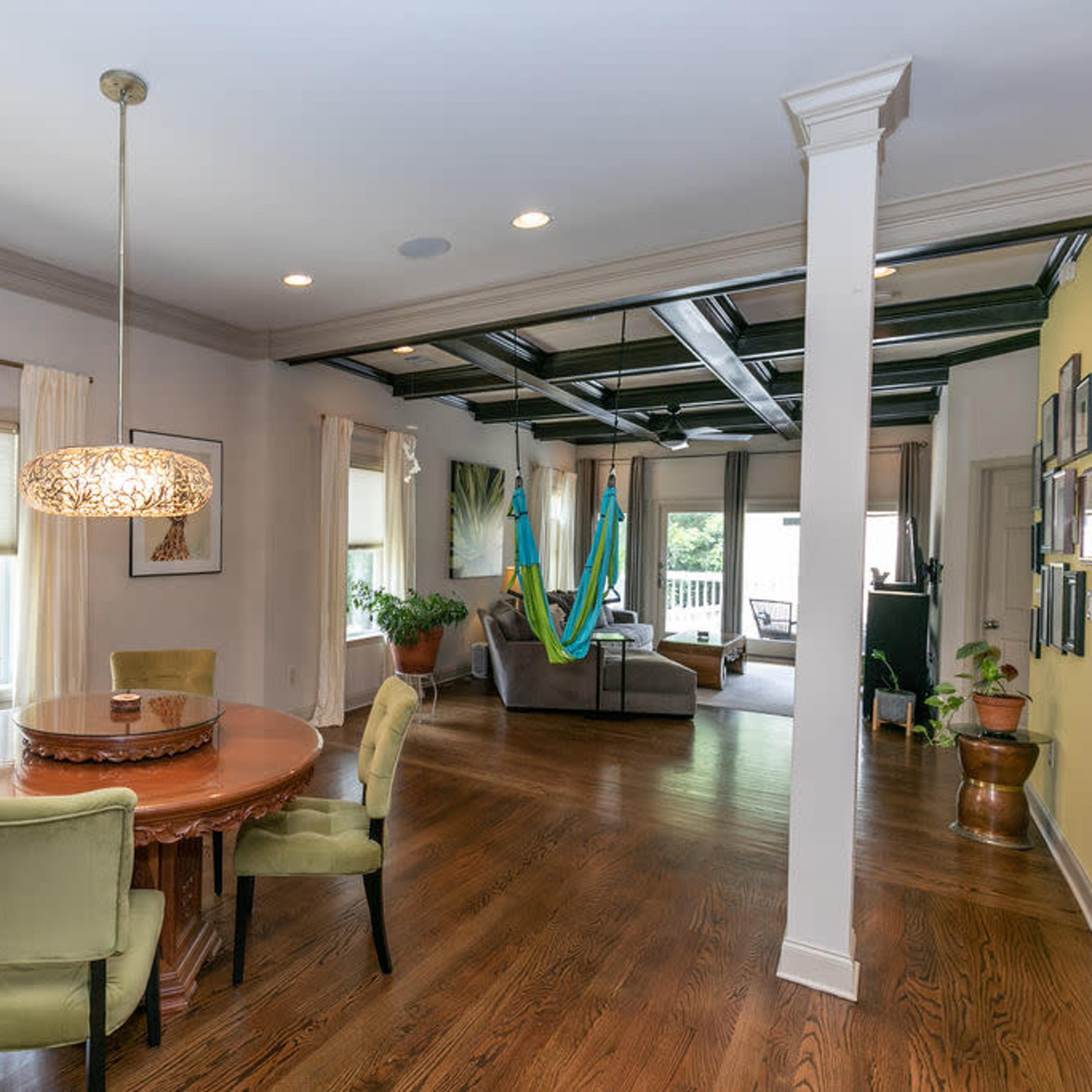A spacious living area with a round dining table, four green chairs, and a view of a sunlit room featuring multiple hammocks and large windows.