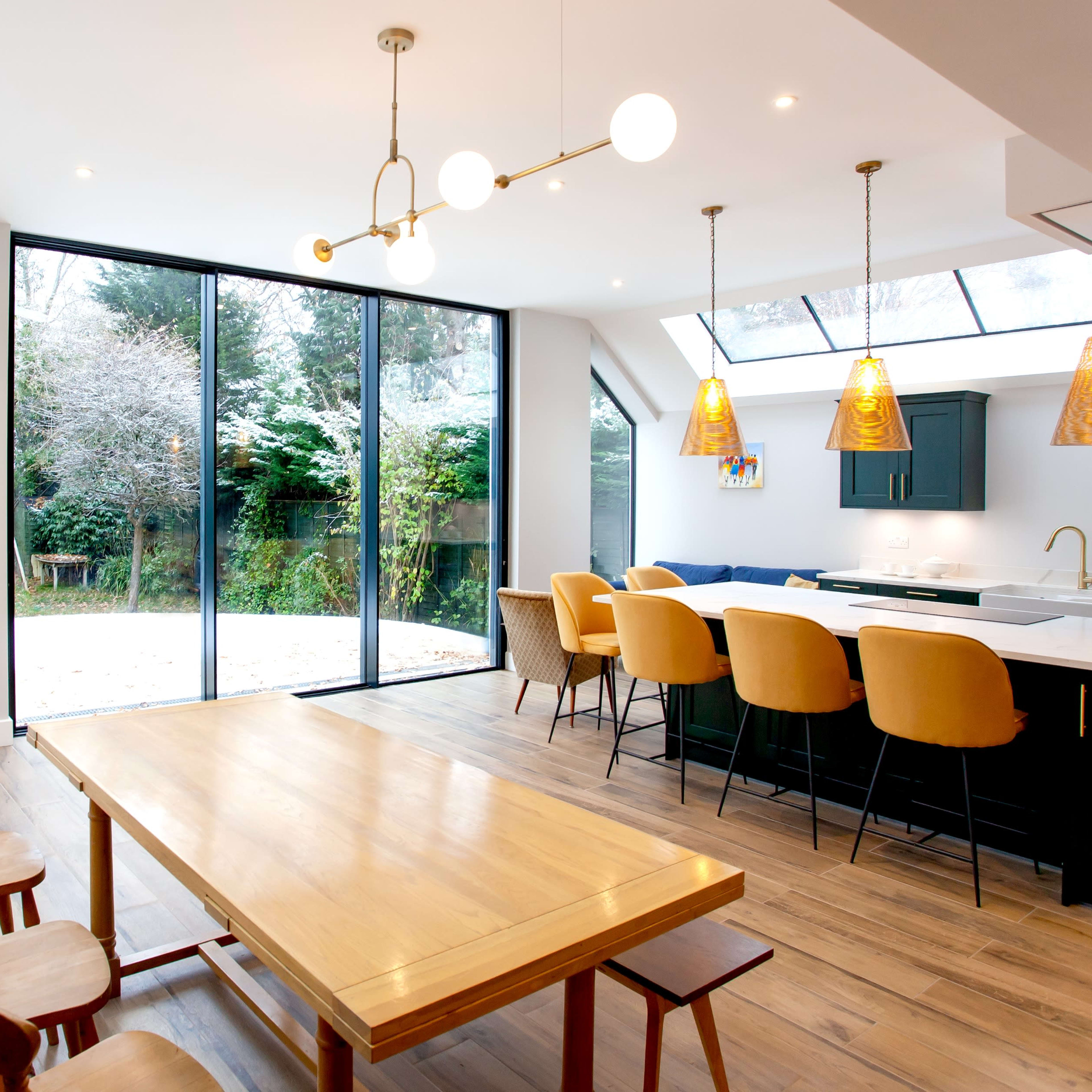 A modern kitchen-dining area features a large wooden table, bar seating, and large glass doors leading to a garden.