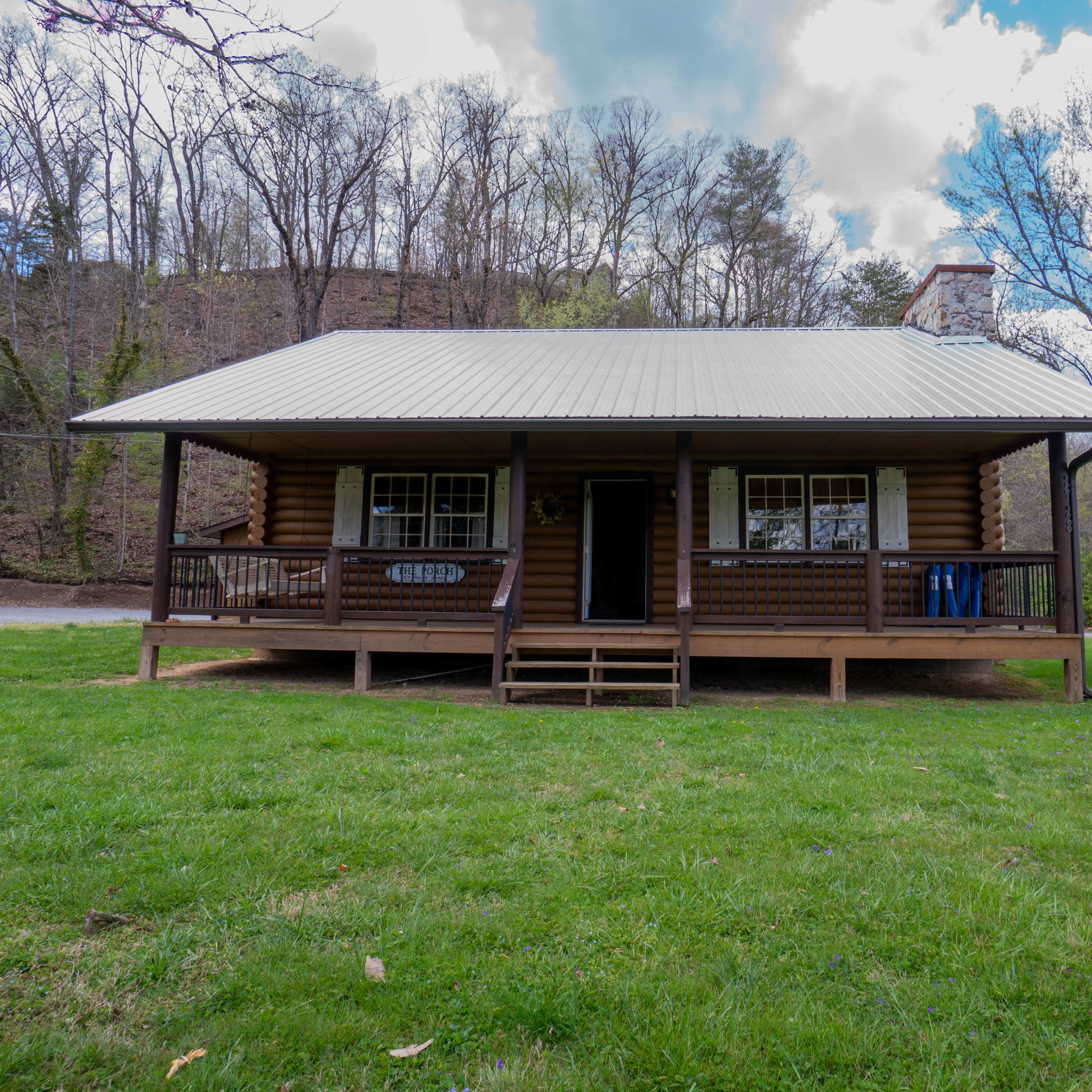 A log cabin with a metal roof features a front porch and is surrounded by green grass and trees.