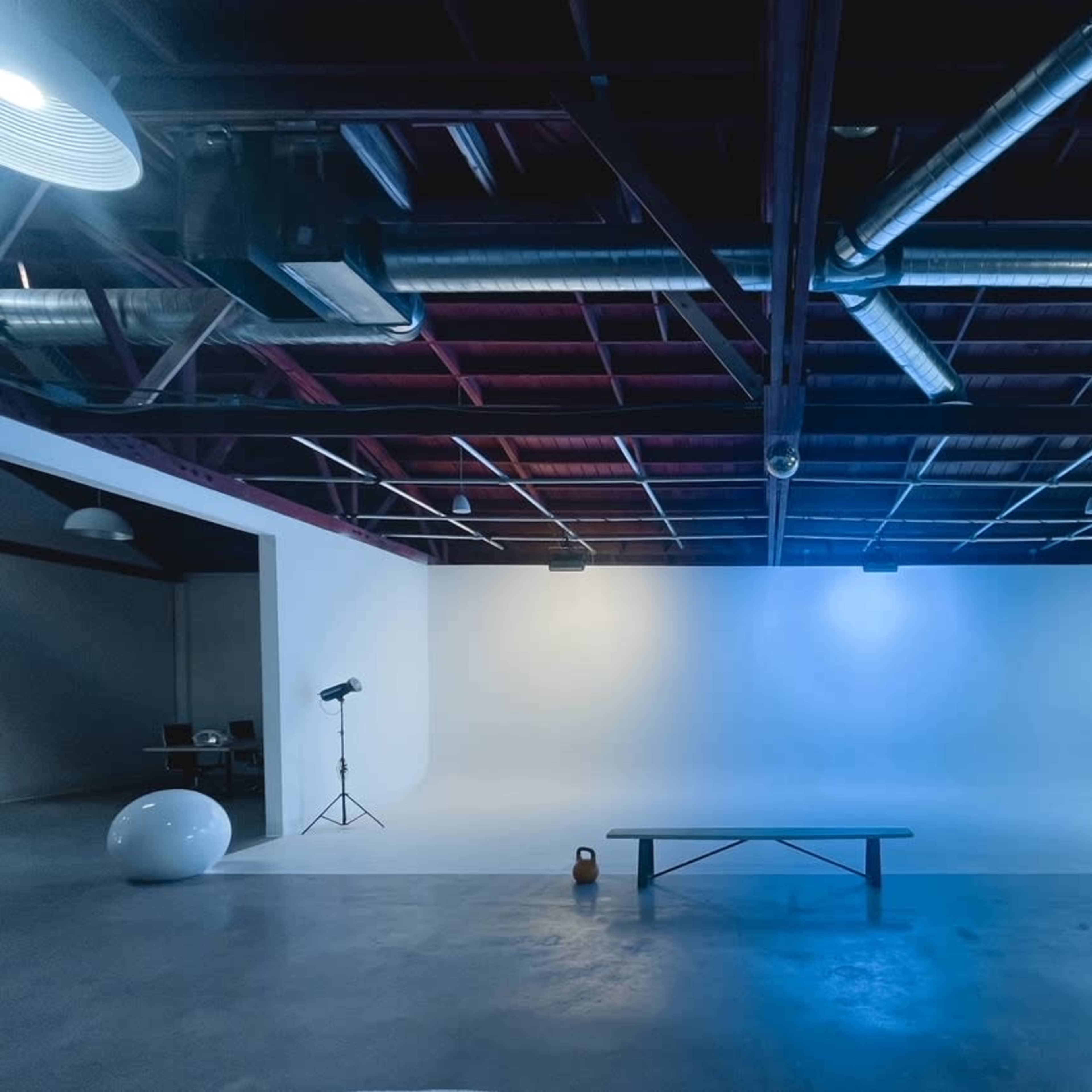A spacious studio space with a curved white backdrop, a wooden bench in the center, and soft lighting from overhead fixtures.