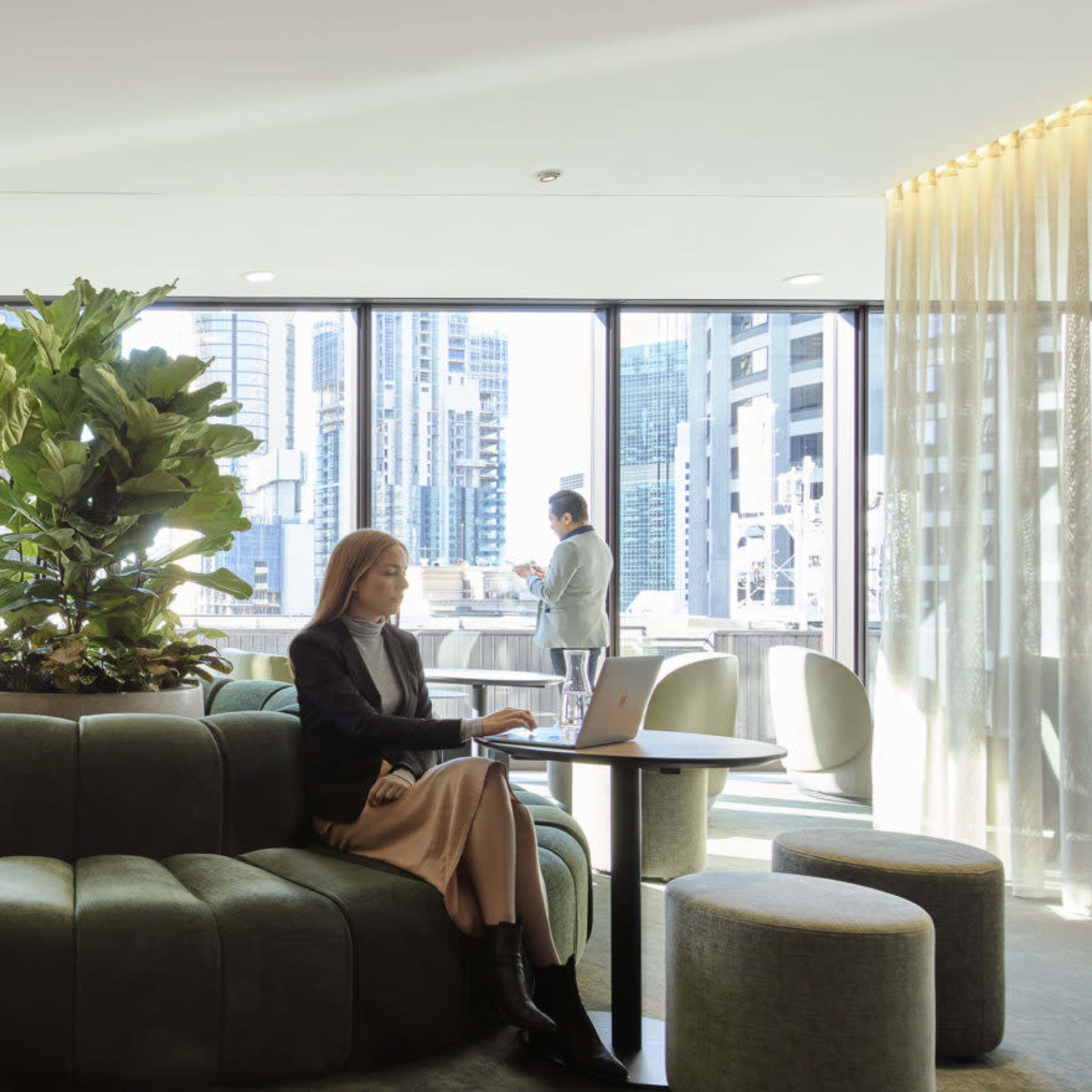 A modern workspace with a circular green seating area, large windows featuring a cityscape, and individuals working on laptops.