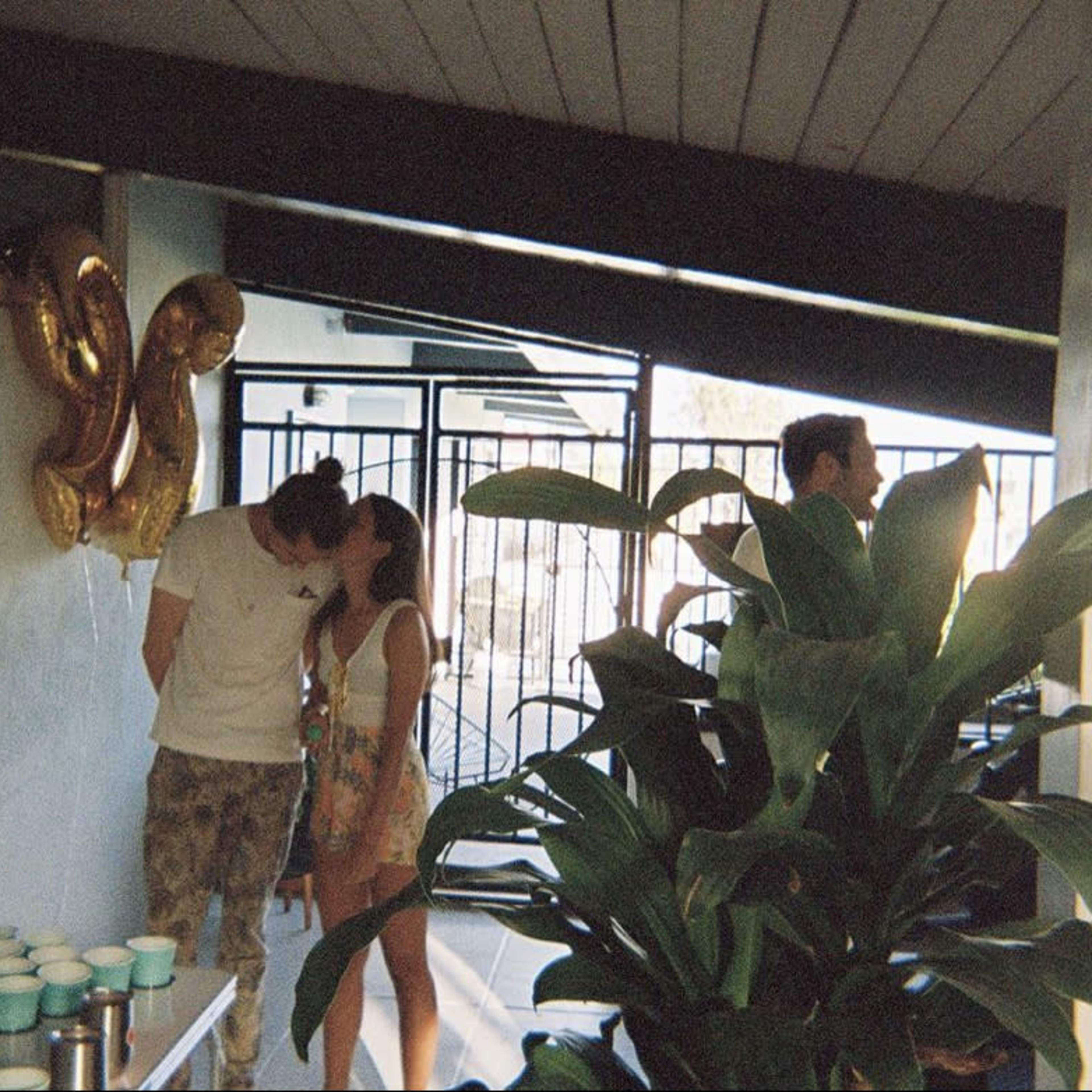 Two people kiss in a room decorated with balloons, while two others stand nearby, and a potted plant is visible in the foreground.