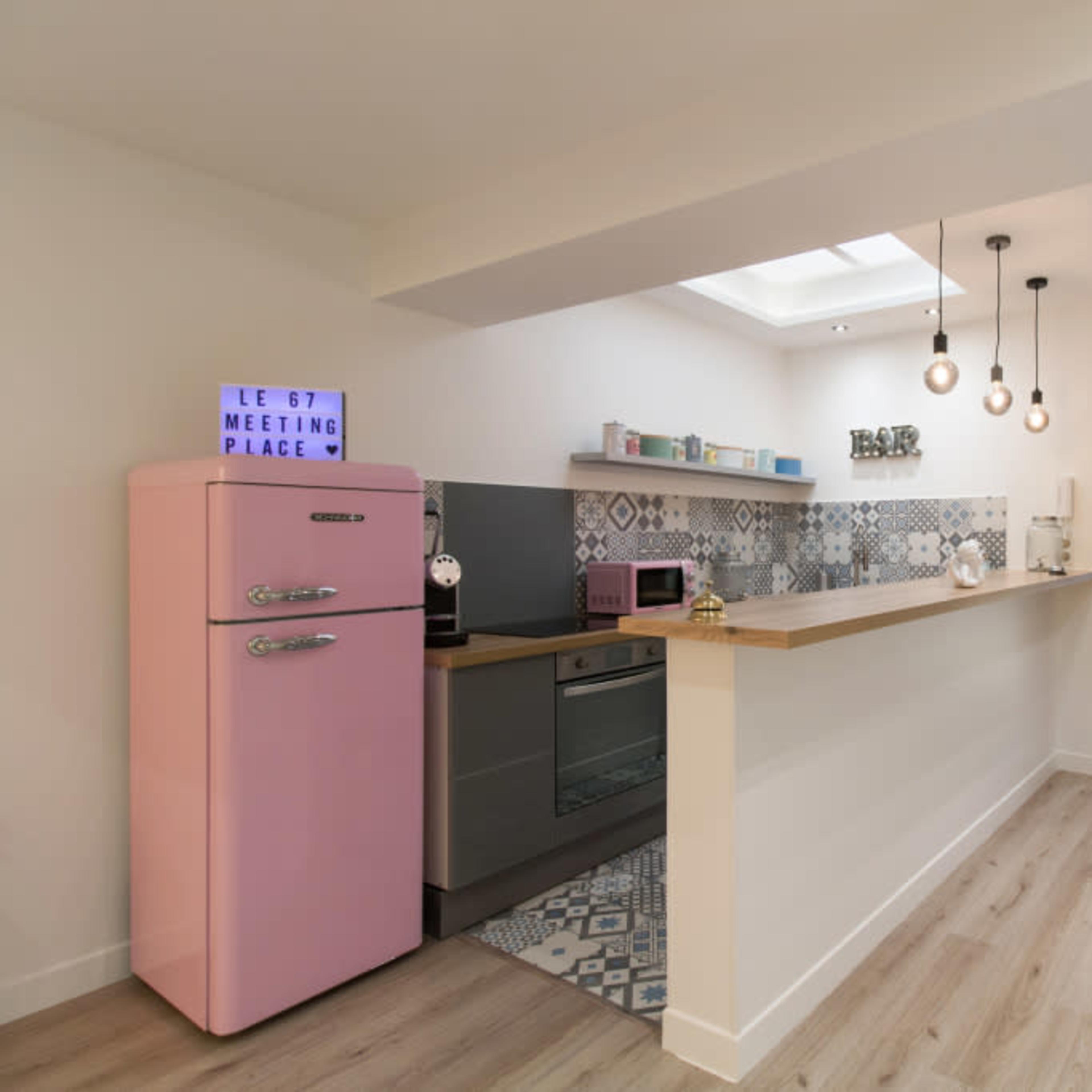 A modern kitchen with a pink refrigerator, a countertop, and decorative tiles, leading to a well-lit hallway with stairs.