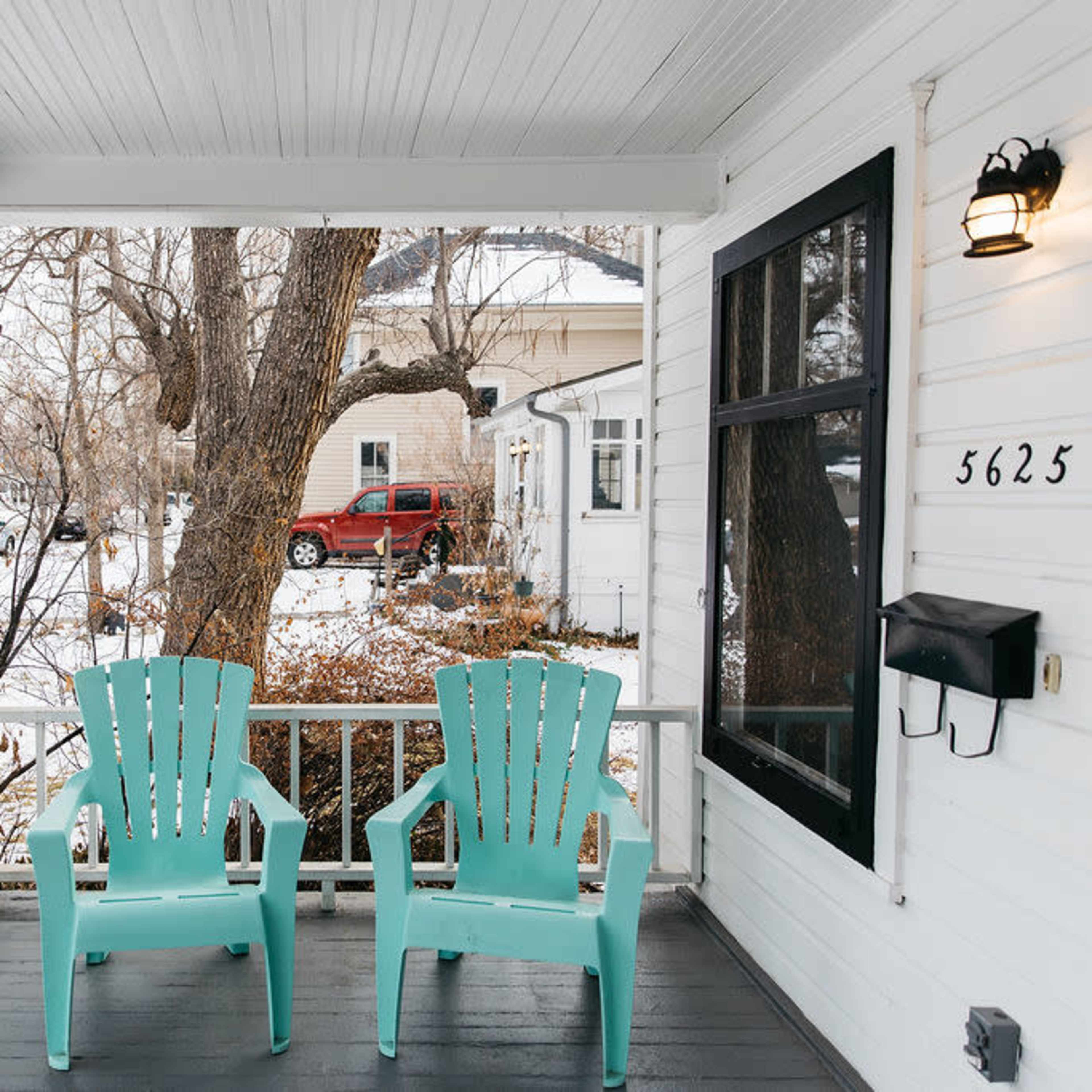 Two turquoise adirondack chairs on a porch facing a snowy front yard with a driveway and nearby houses.