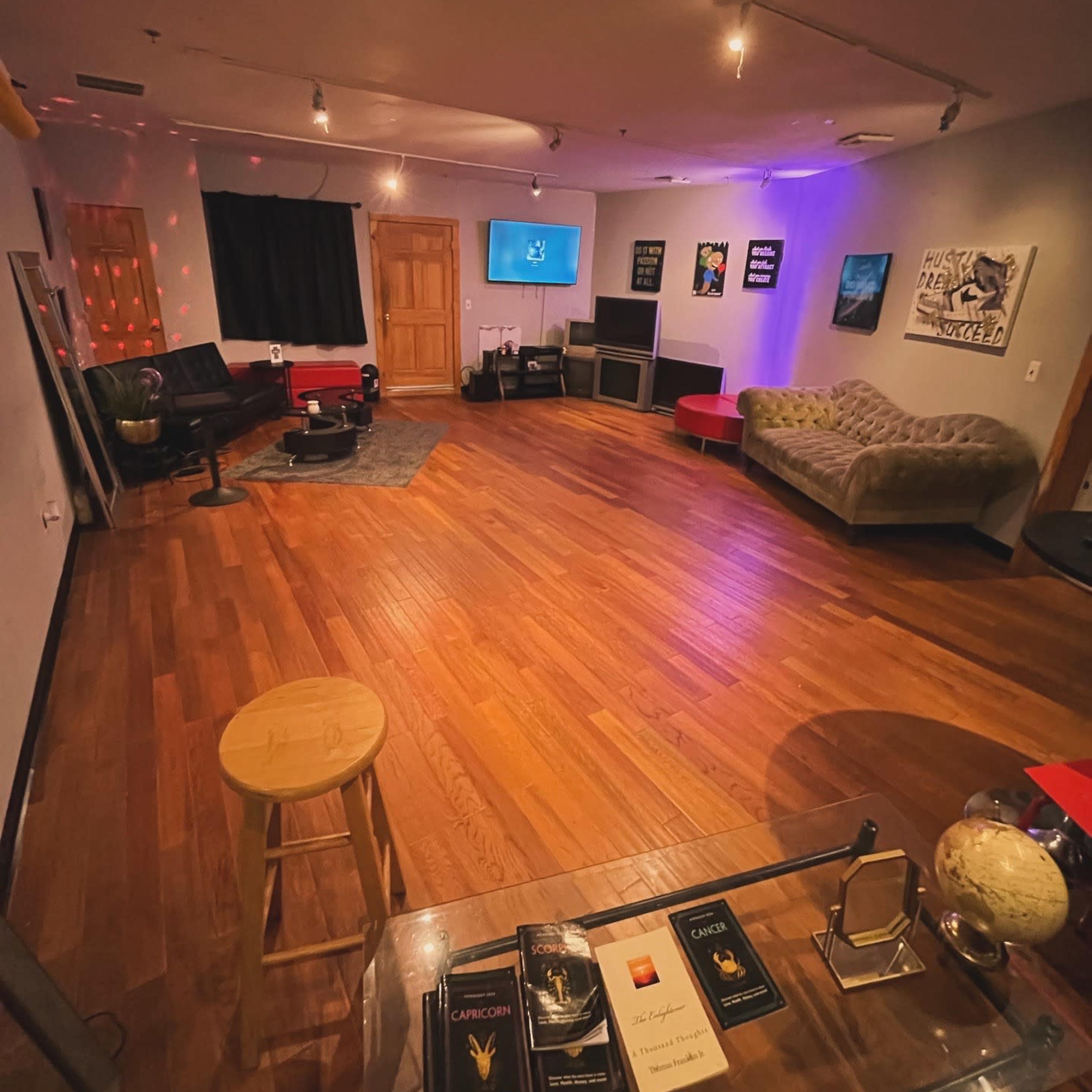 A spacious, empty room with wooden flooring, a few pieces of furniture including a couch and chairs, and a television on the wall.