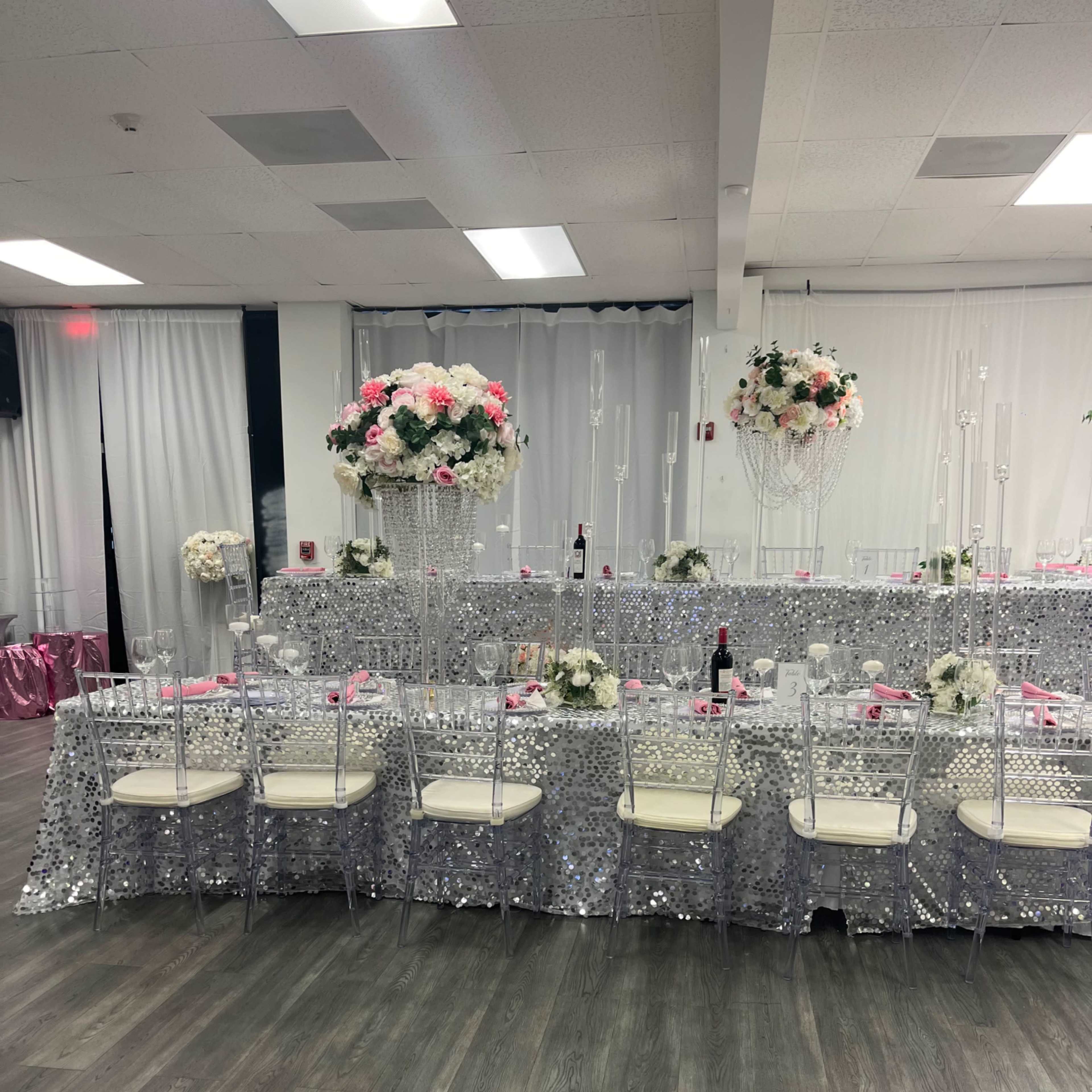 A banquet hall setup with several tables covered in silver sequin tablecloths, adorned with floral centerpieces and white chairs arranged around them.