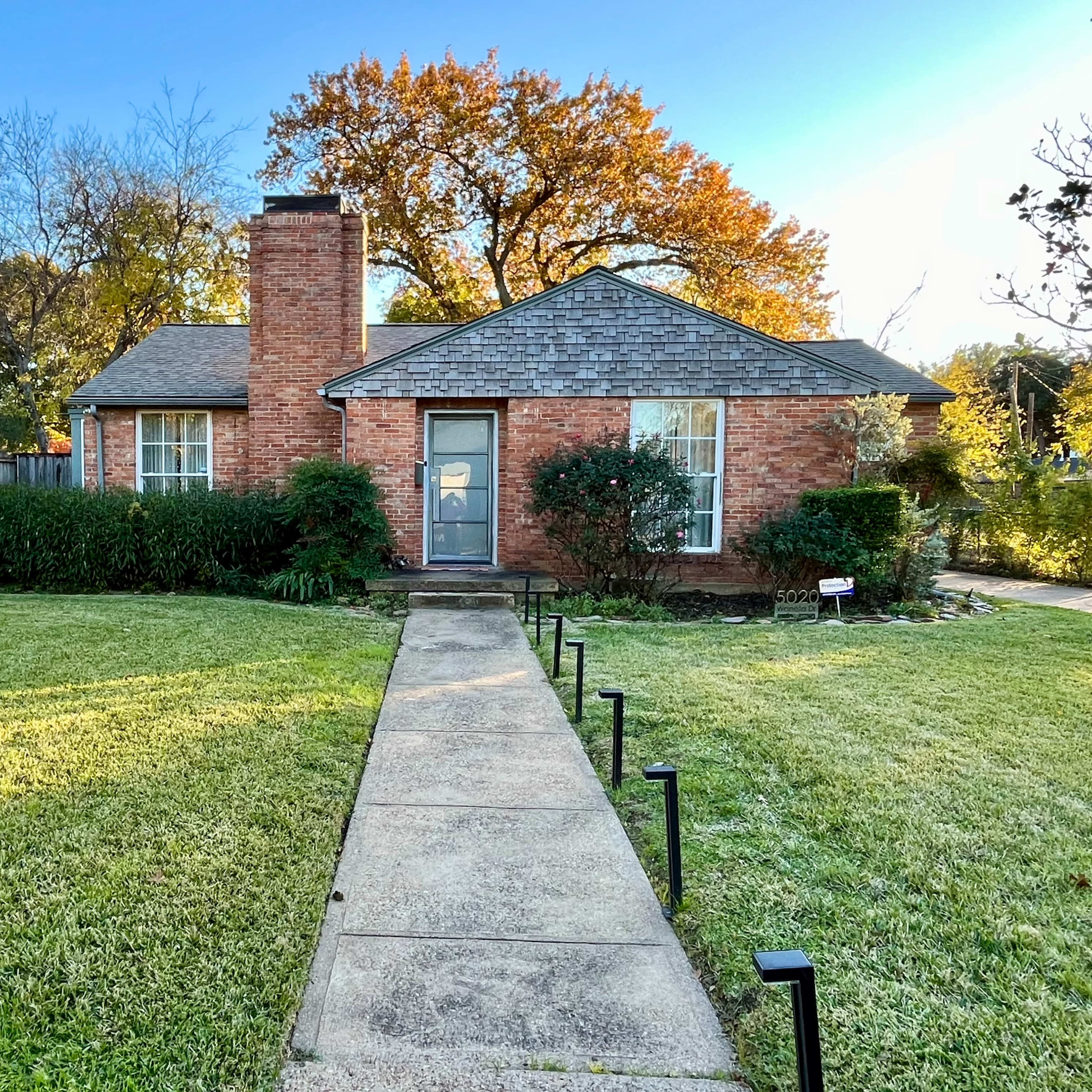 A brick house with a chimney and a front path is surrounded by a well-maintained lawn and shrubs.