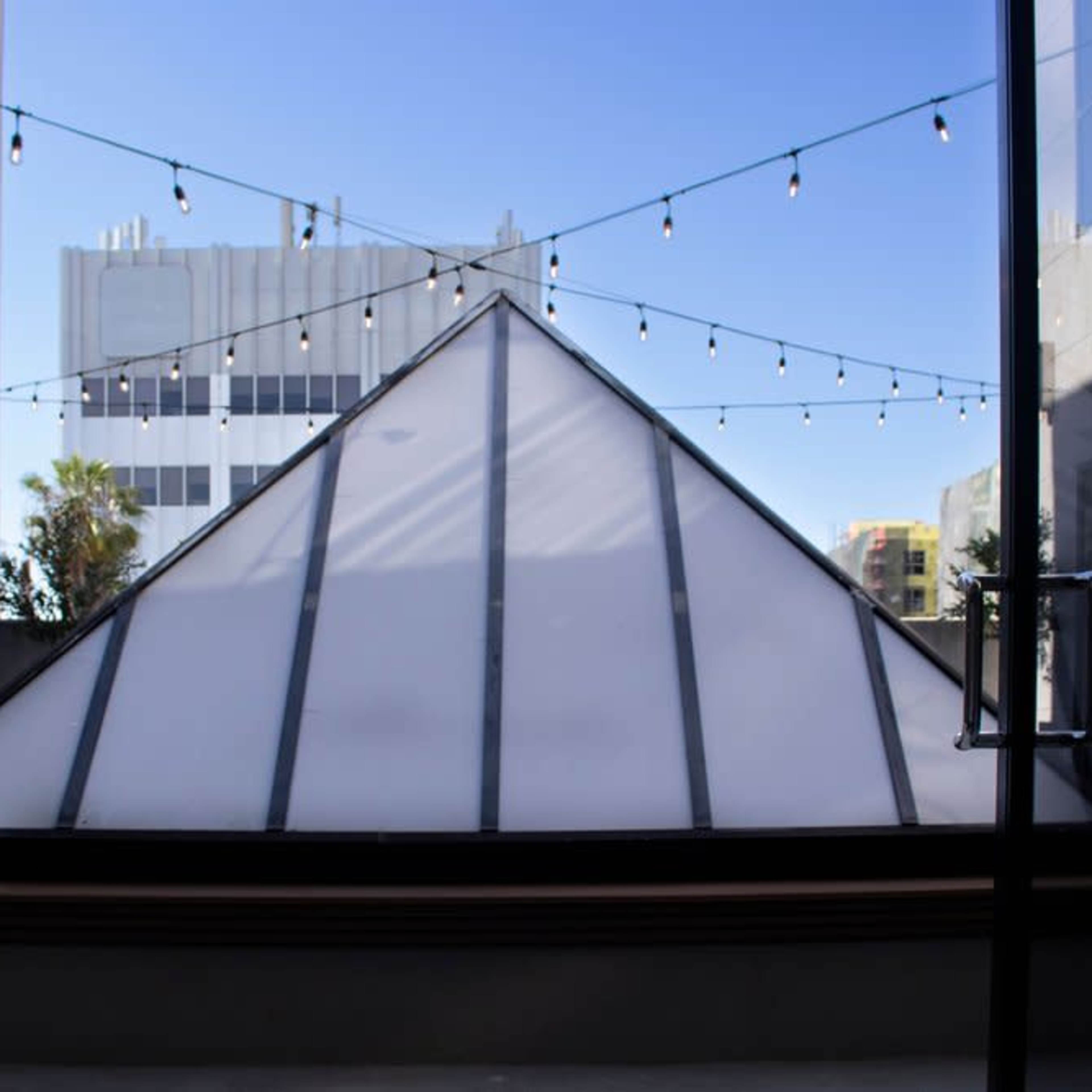 A large pyramid-shaped structure made of glass is positioned in an outdoor area between two buildings, with string lights overhead and a clear blue sky in the background.