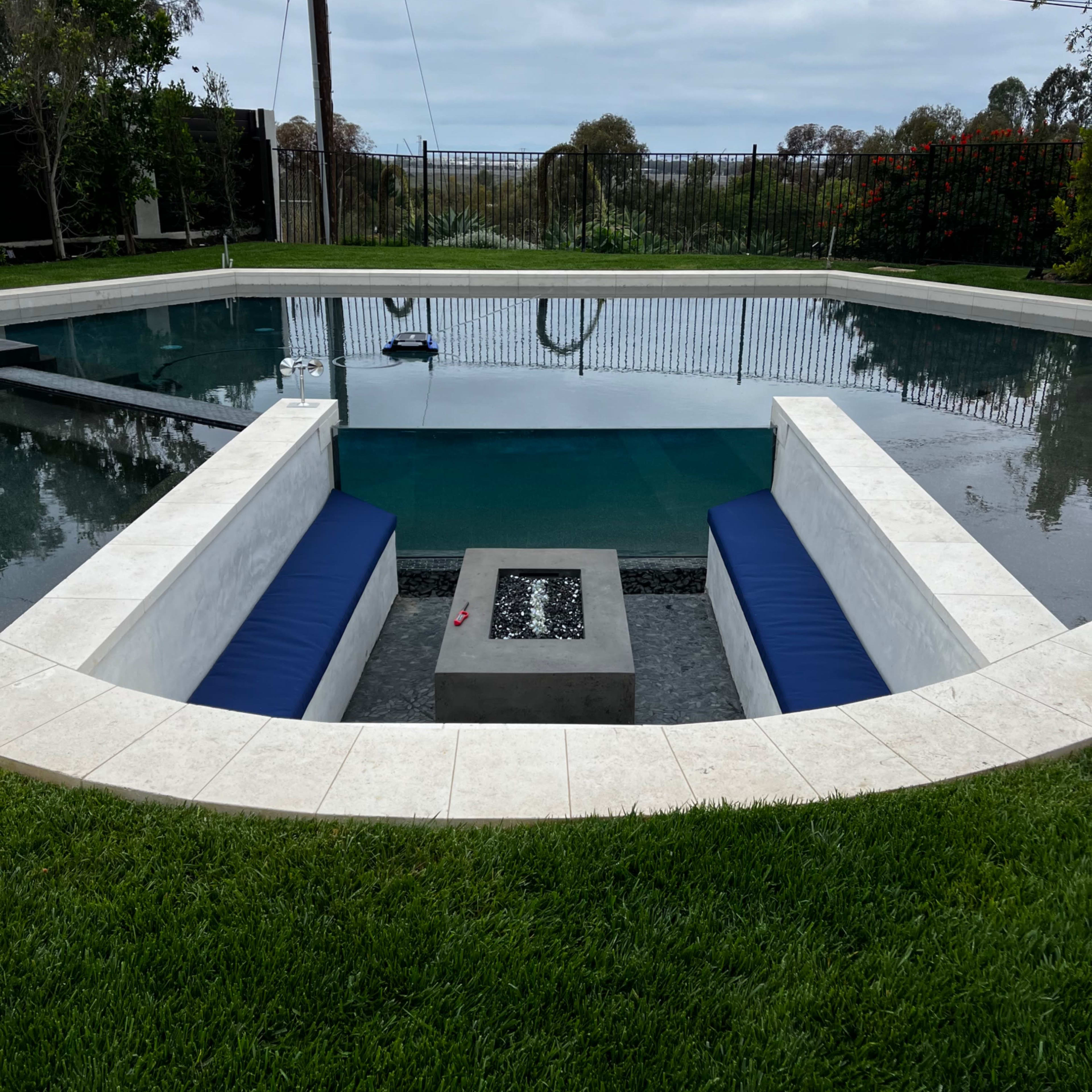 A rectangular swimming pool with a sunken seating area at one end, surrounded by grass and a fencing barrier.