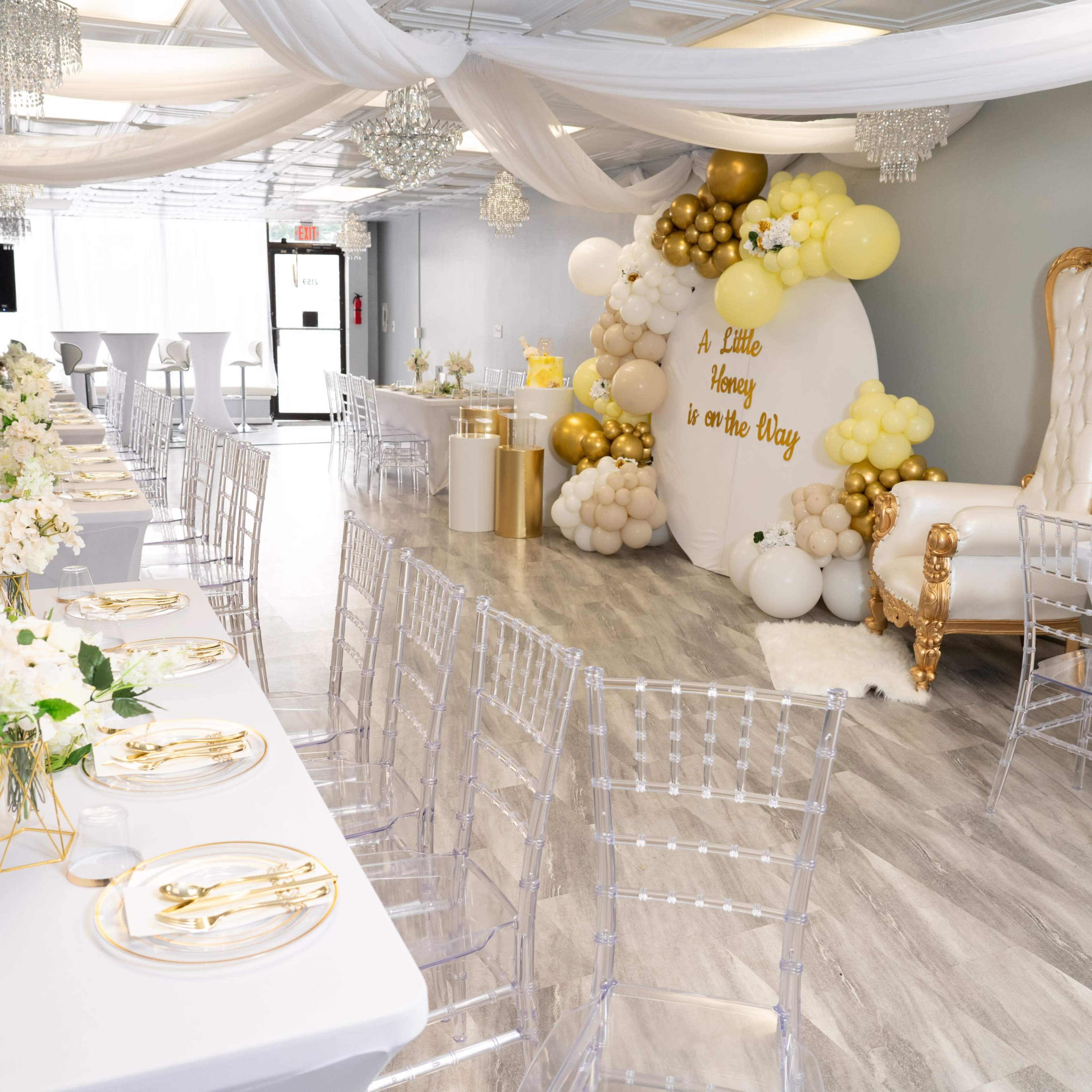 A decorated event space with white tables set with gold accents, a backdrop with balloons, and an ornate throne chair.