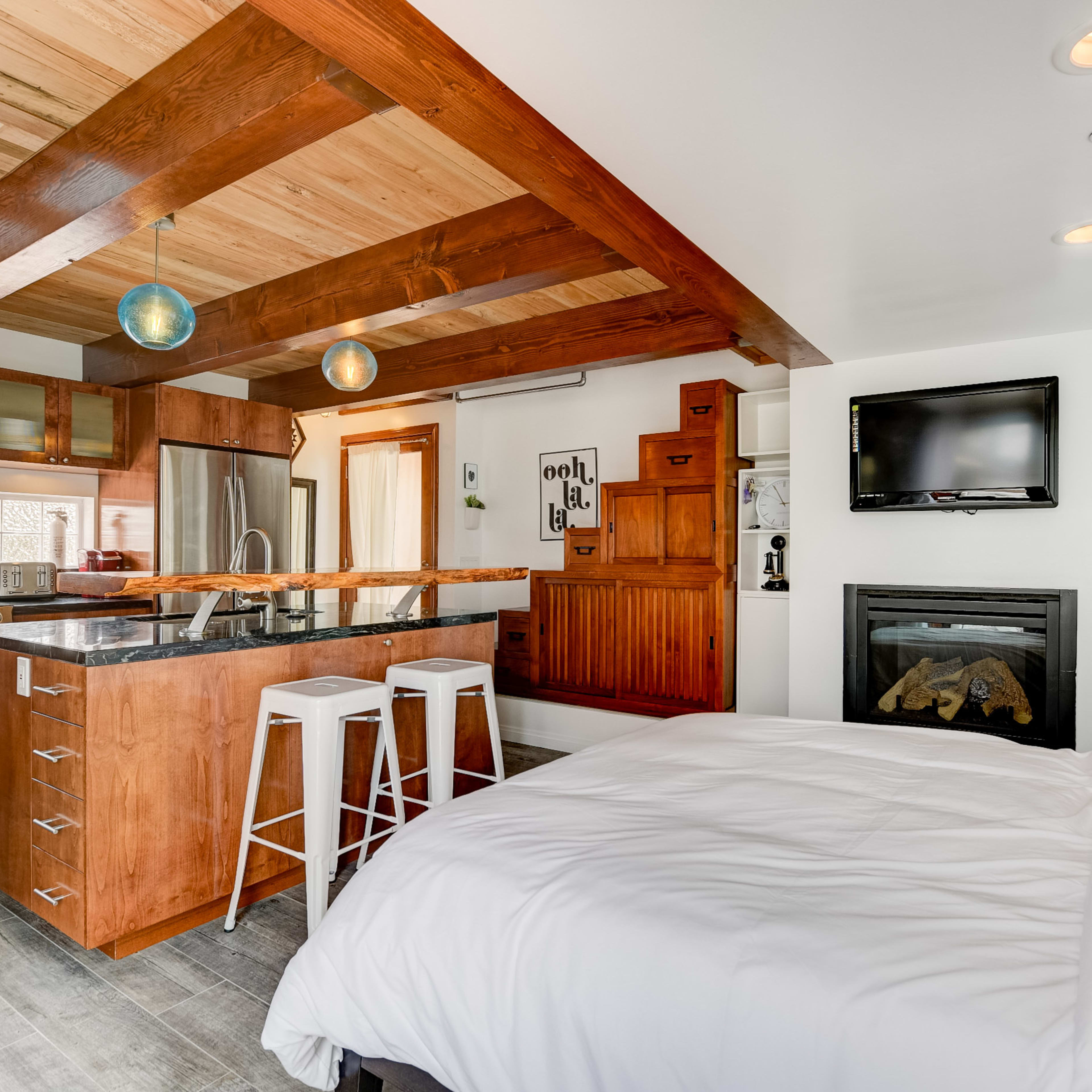 A compact studio apartment featuring a kitchen with wooden beams, a small dining area with stools, a bed, and a fireplace.