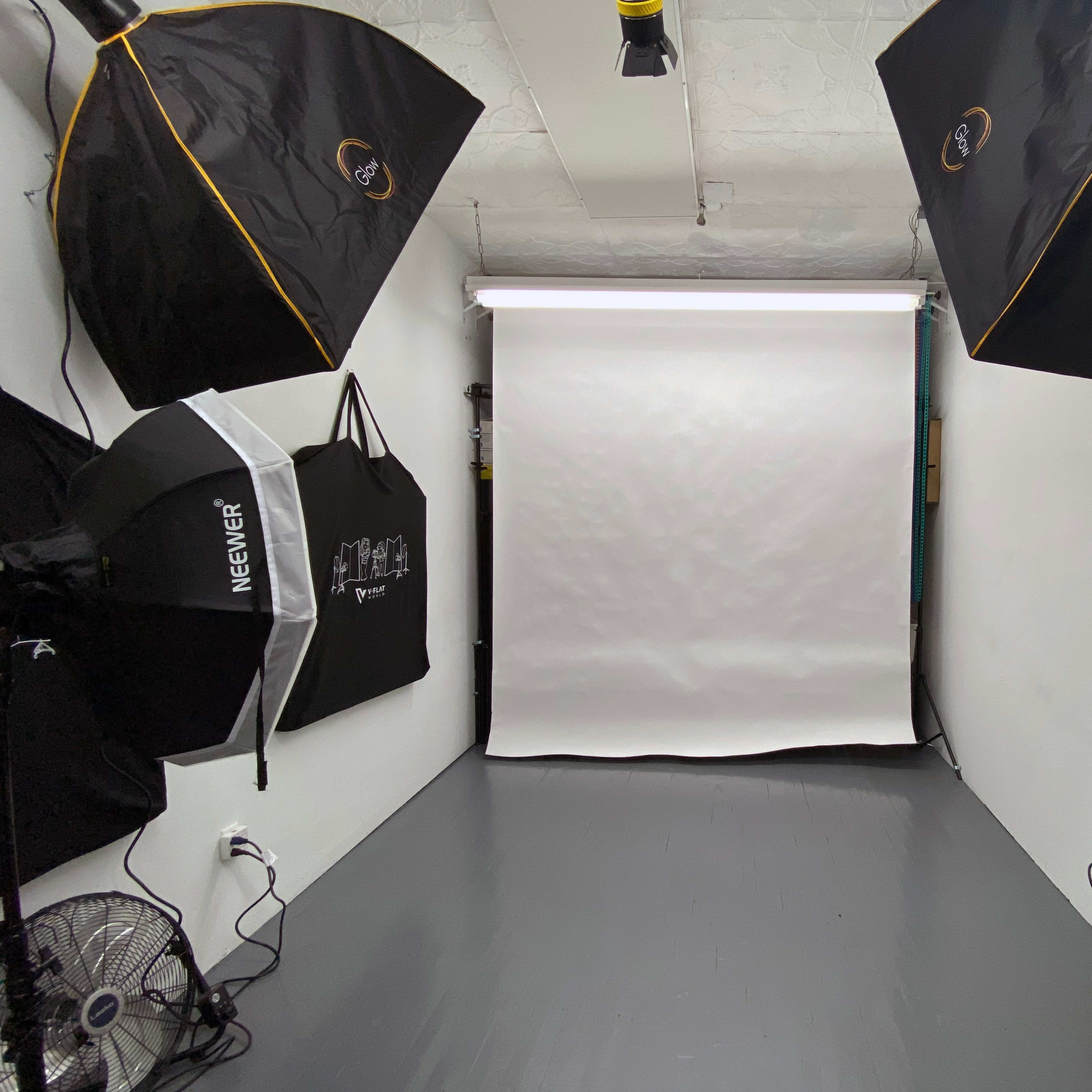 A photography studio with two large softbox lights, a white backdrop, and a fan on the floor.