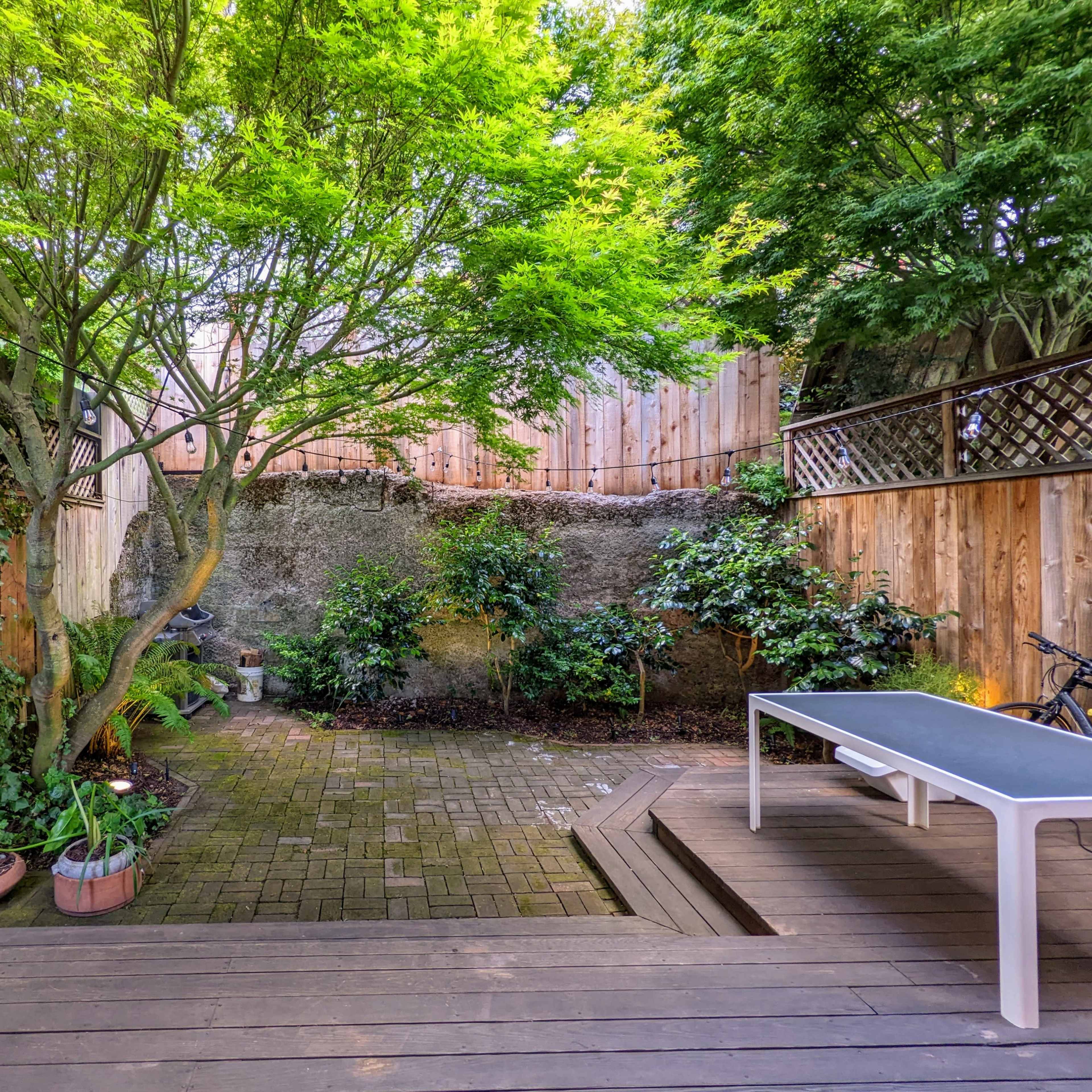 A fenced backyard features a stone-paved area surrounded by greenery and plants, with a white table and a bicycle parked nearby.