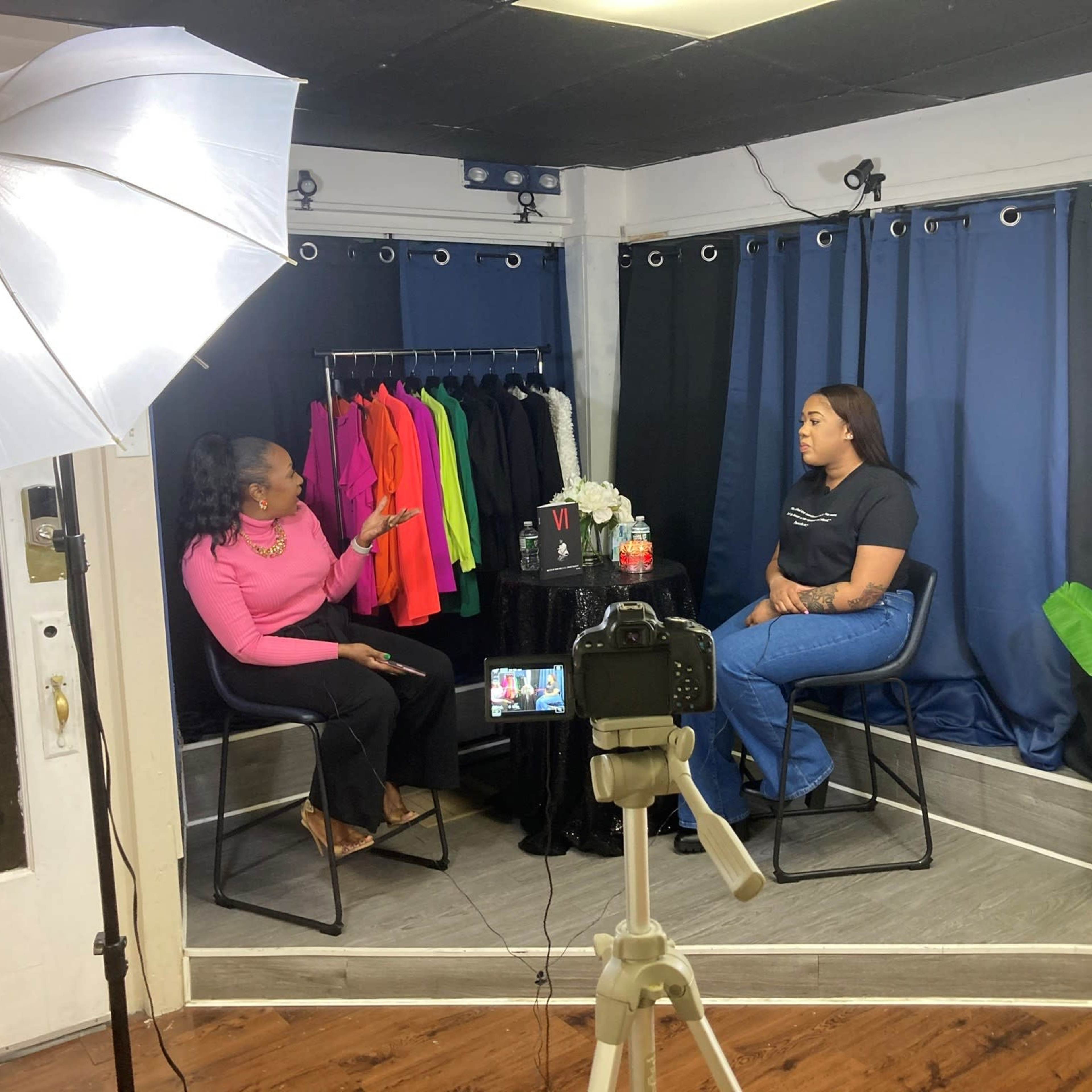 Two women are seated in a studio setting for an interview, with a rack of colorful clothing visible in the background.