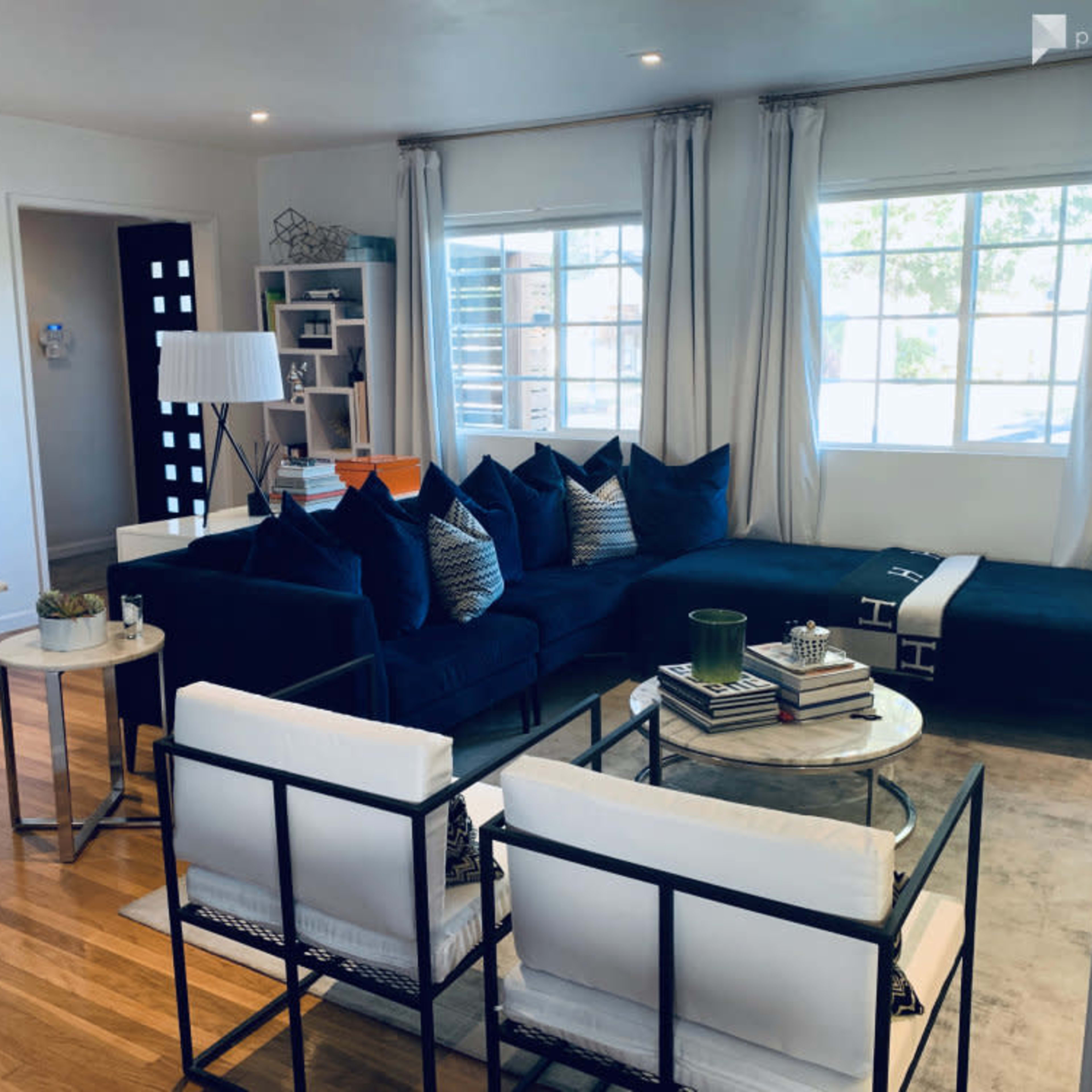 A modern living room featuring a dark blue sectional sofa, two white chairs, a coffee table, and large windows allowing natural light.
