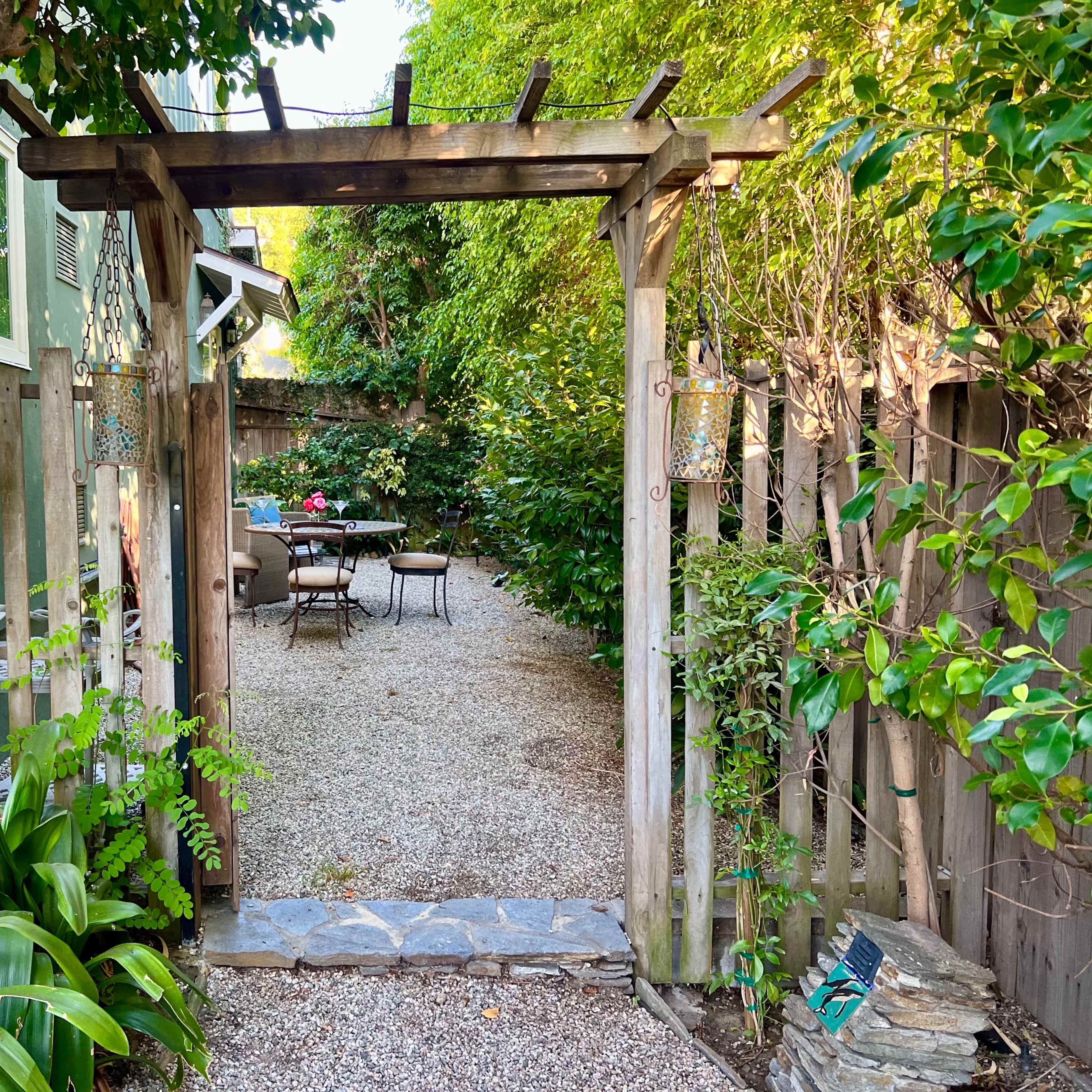 A gravel pathway leads through a wooden archway into a garden space with seating surrounded by greenery.