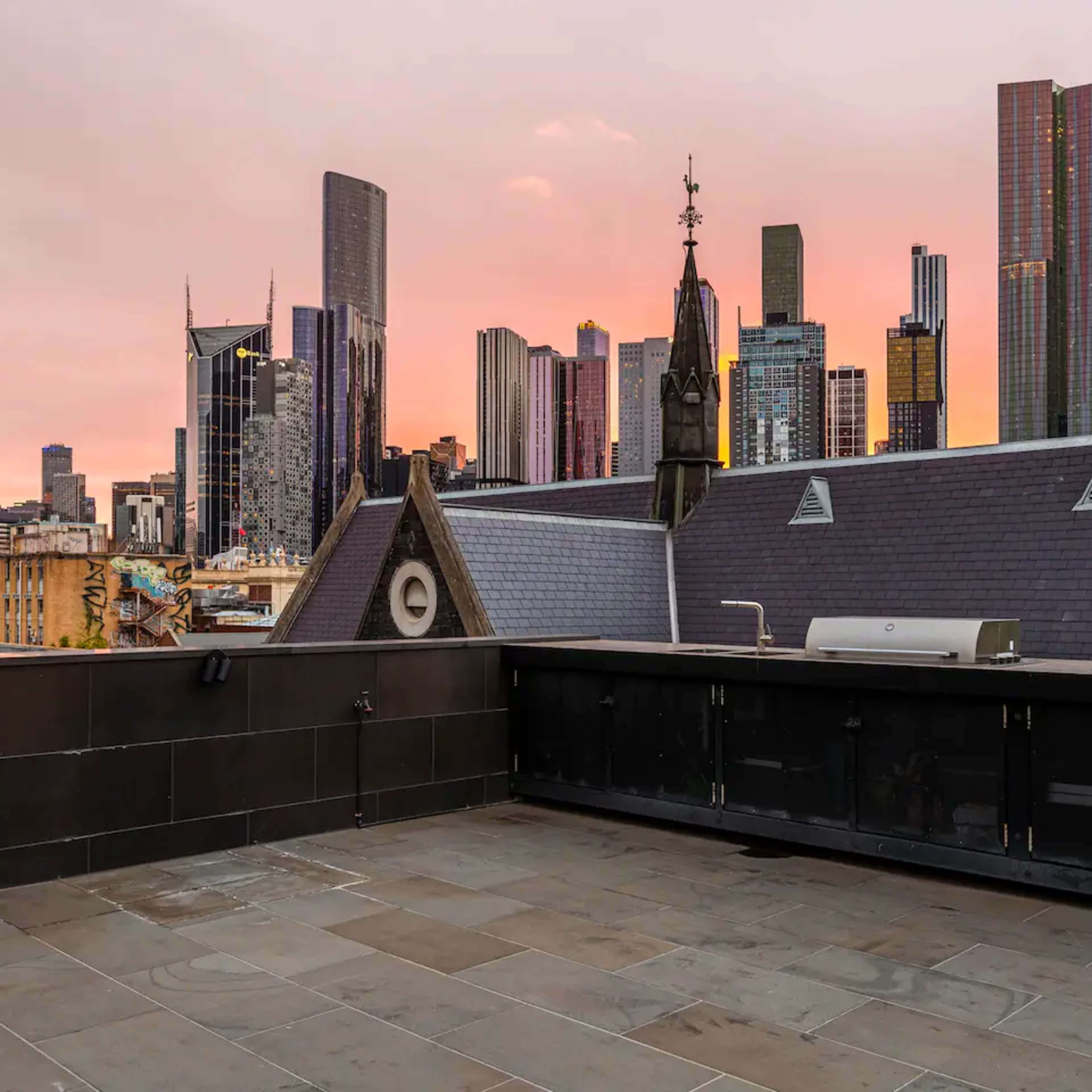 A rooftop terrace with a view of a city skyline at sunset, featuring modern skyscrapers and a historic building facade.