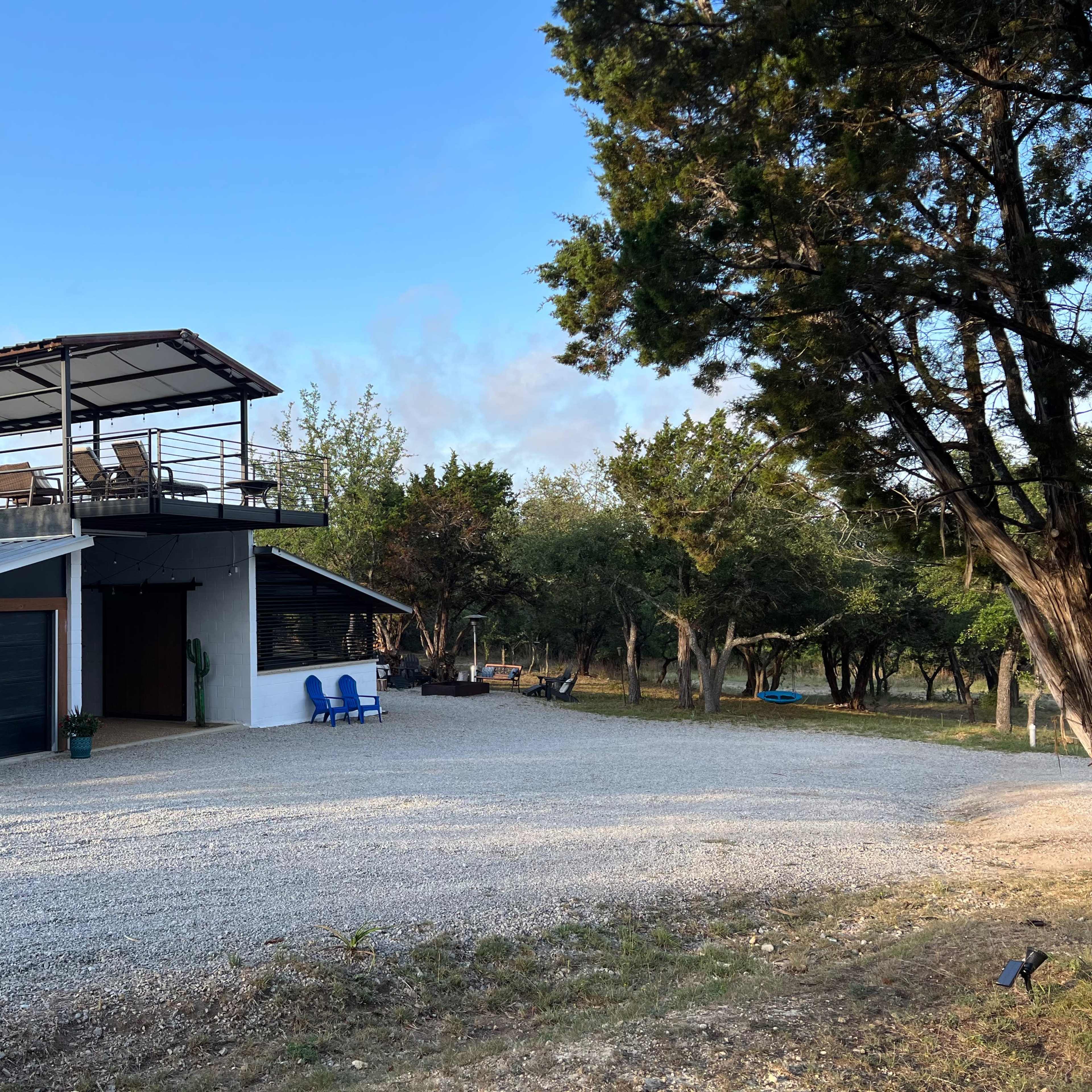 A modern two-story house with a rooftop terrace is located on a gravel driveway surrounded by trees.