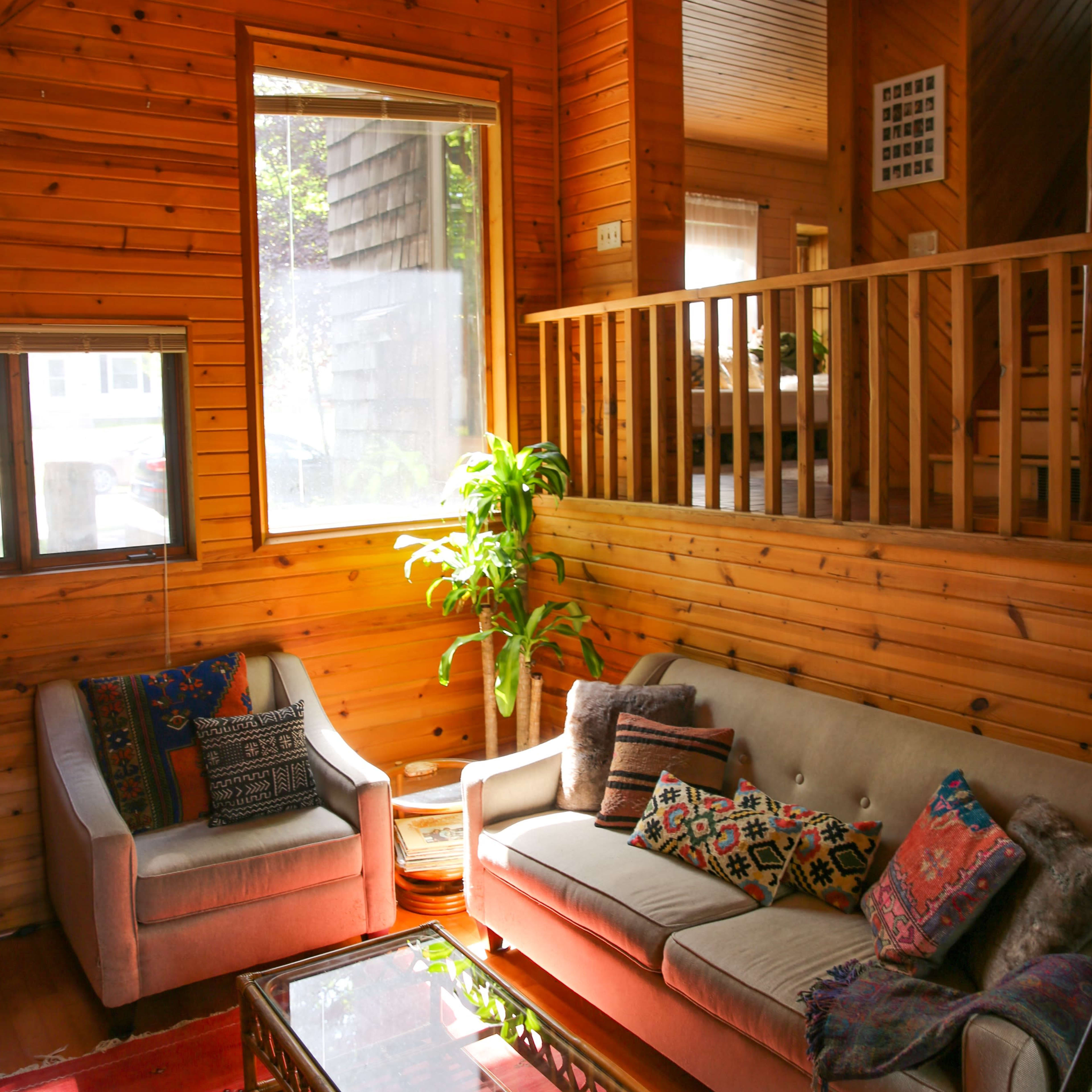 A cozy living room with wooden walls, a gray couch adorned with colorful pillows, a glass coffee table, and a potted plant near a large window.