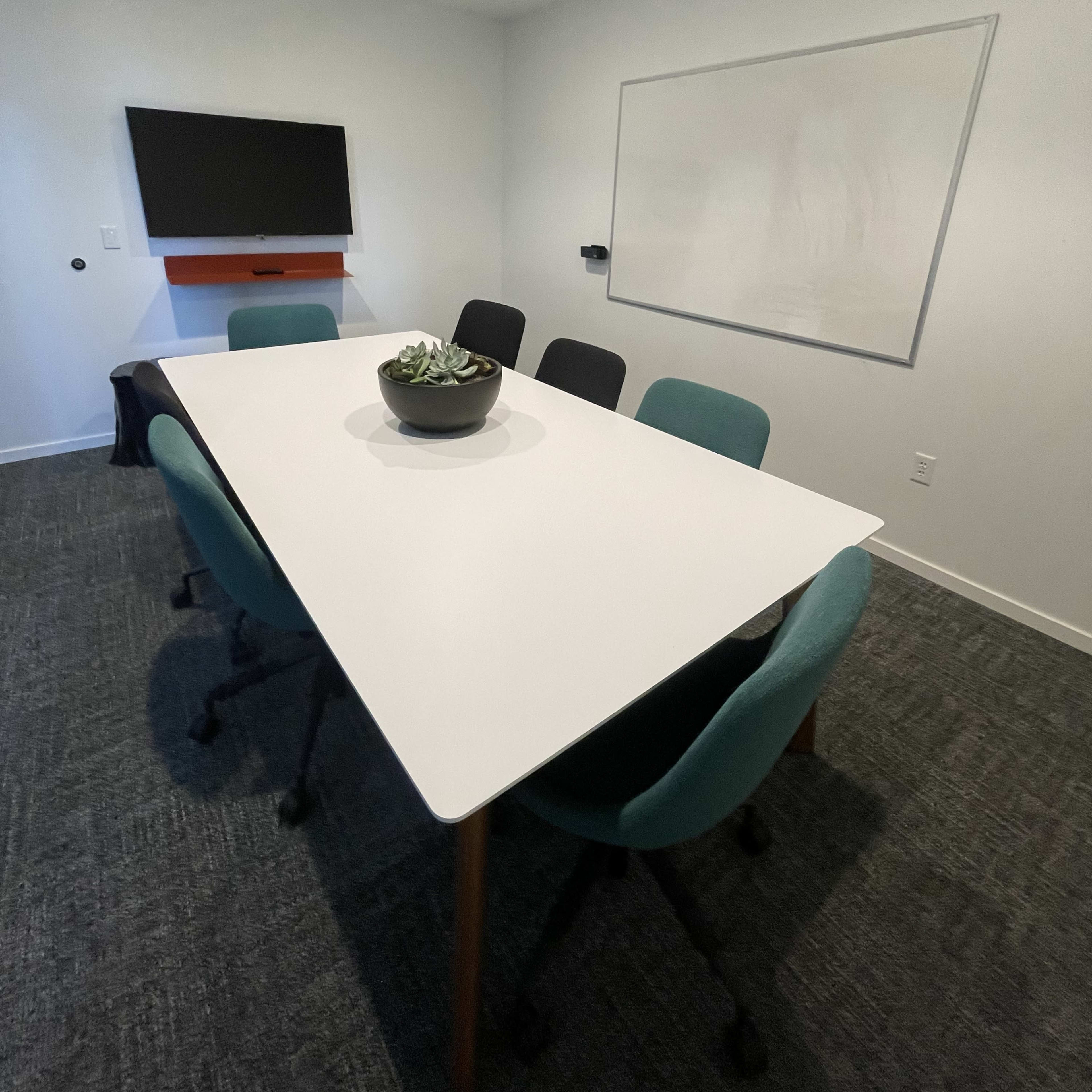 A conference room features a large white table surrounded by chairs, with a TV mounted on the wall and a blank whiteboard.