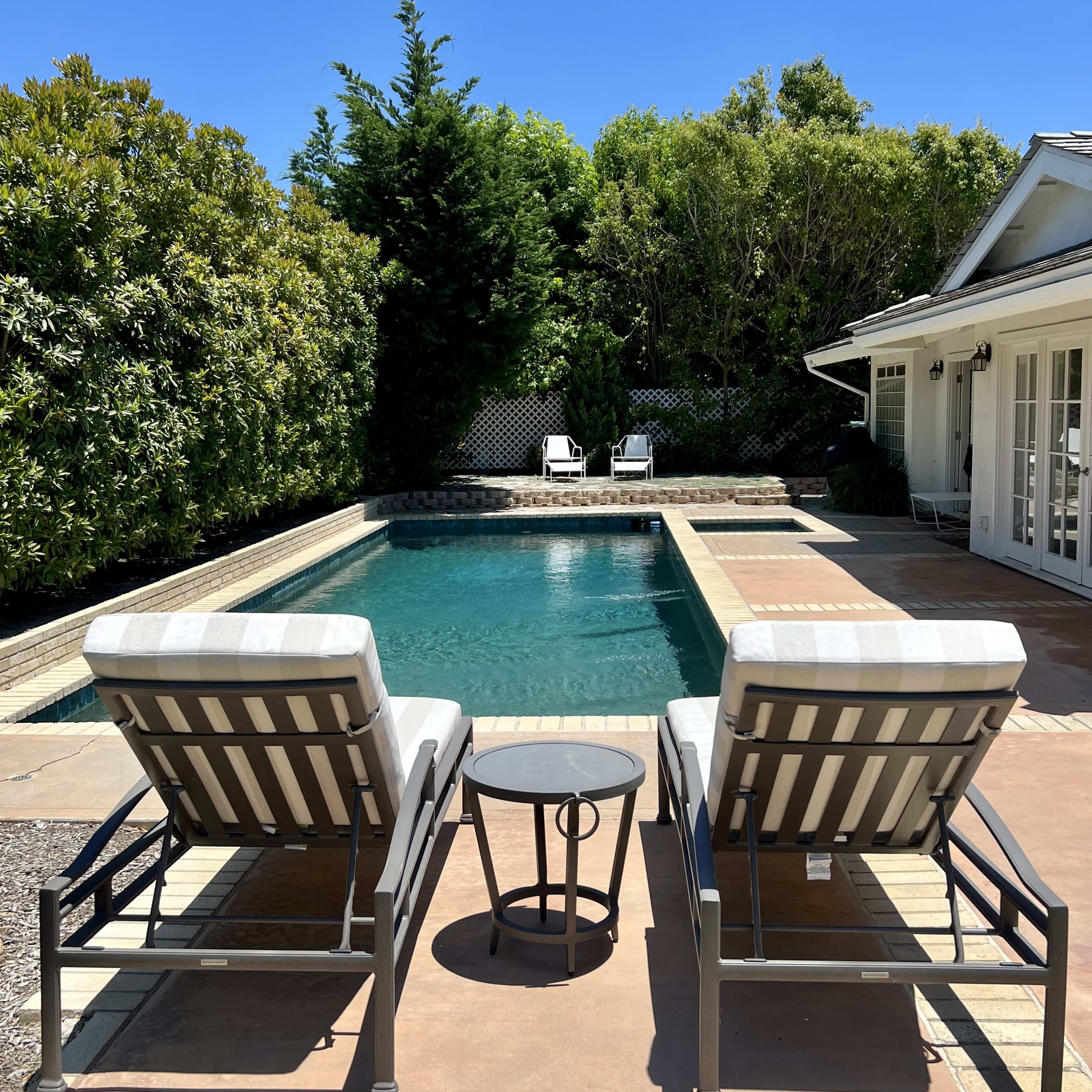 Two reclining lounge chairs flank a small table beside a swimming pool surrounded by green foliage and a sunny blue sky.
