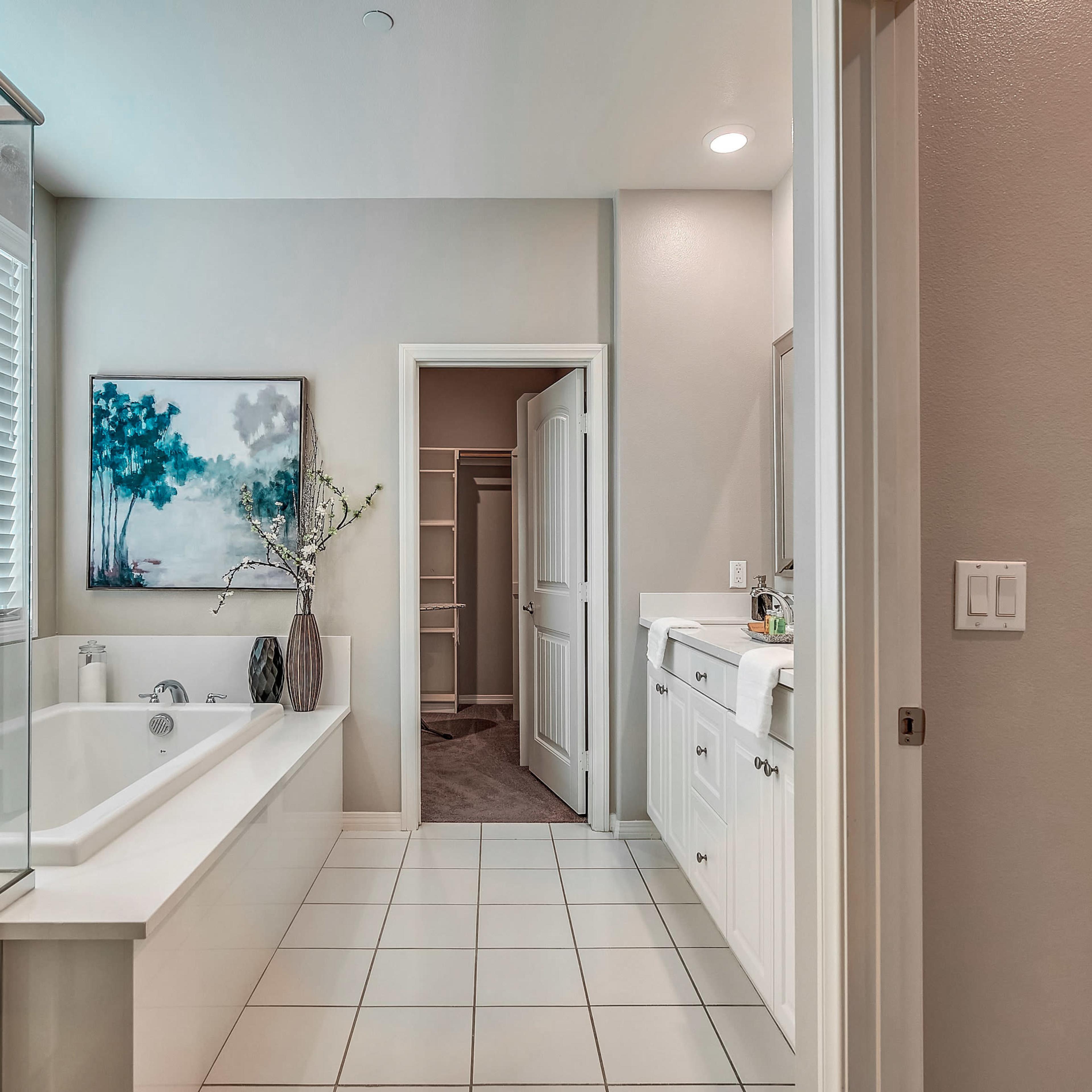 A modern bathroom with a glass shower, a freestanding bathtub, and a vanity area, featuring a painting on the wall and a doorway leading to a closet.