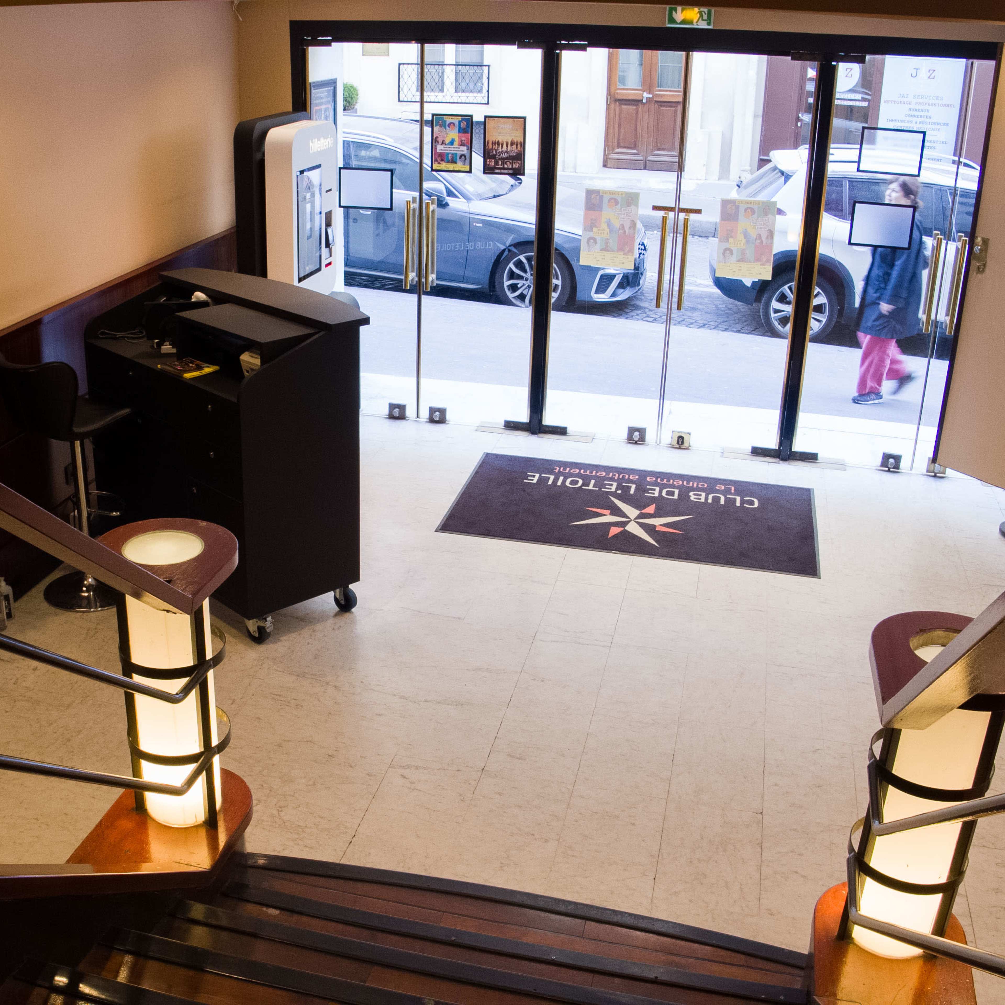 A view from the top of a staircase leading into a lobby area with a reception desk and glass doors opening to the street.