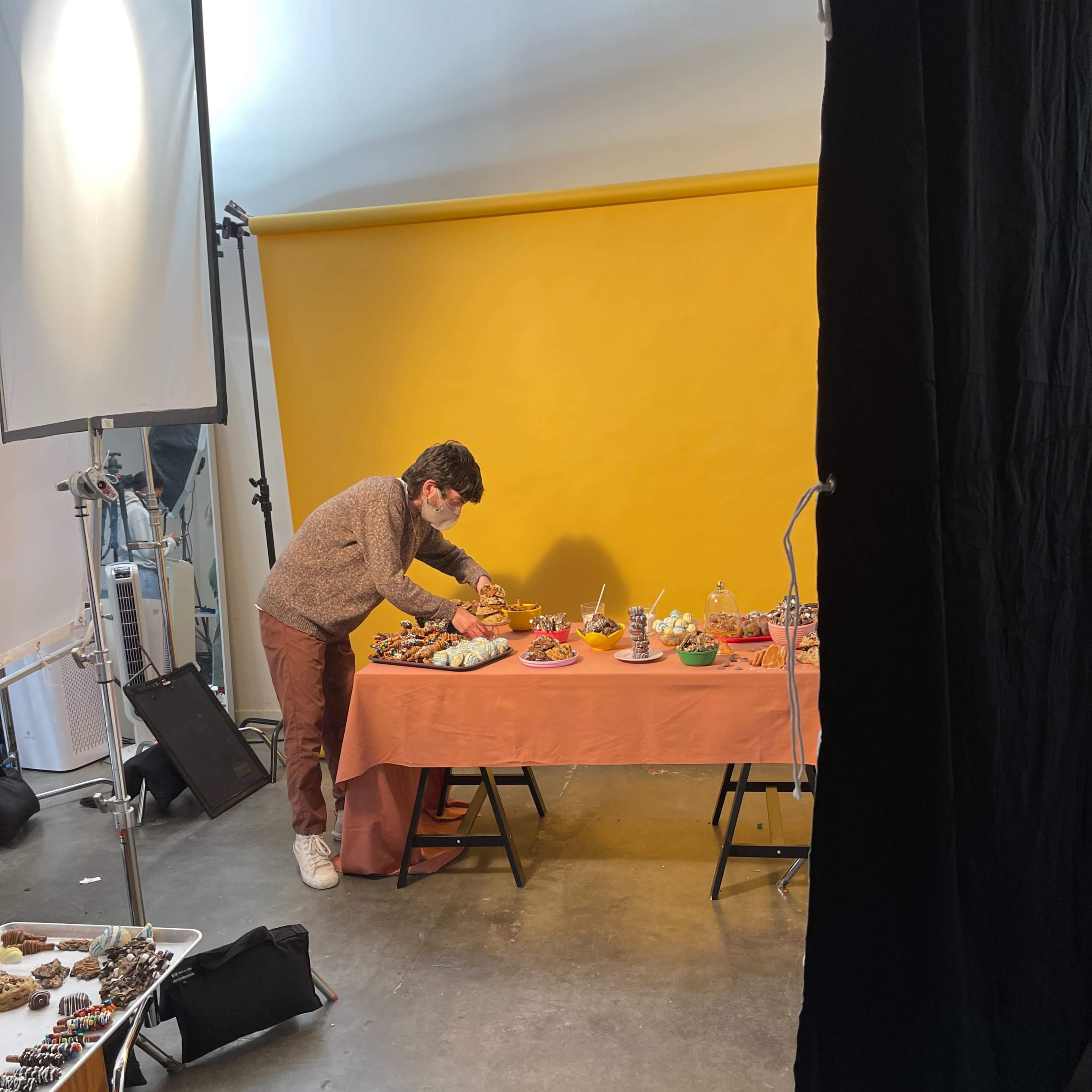 A person is arranging food items on a table in front of a bright yellow backdrop in a studio setting.