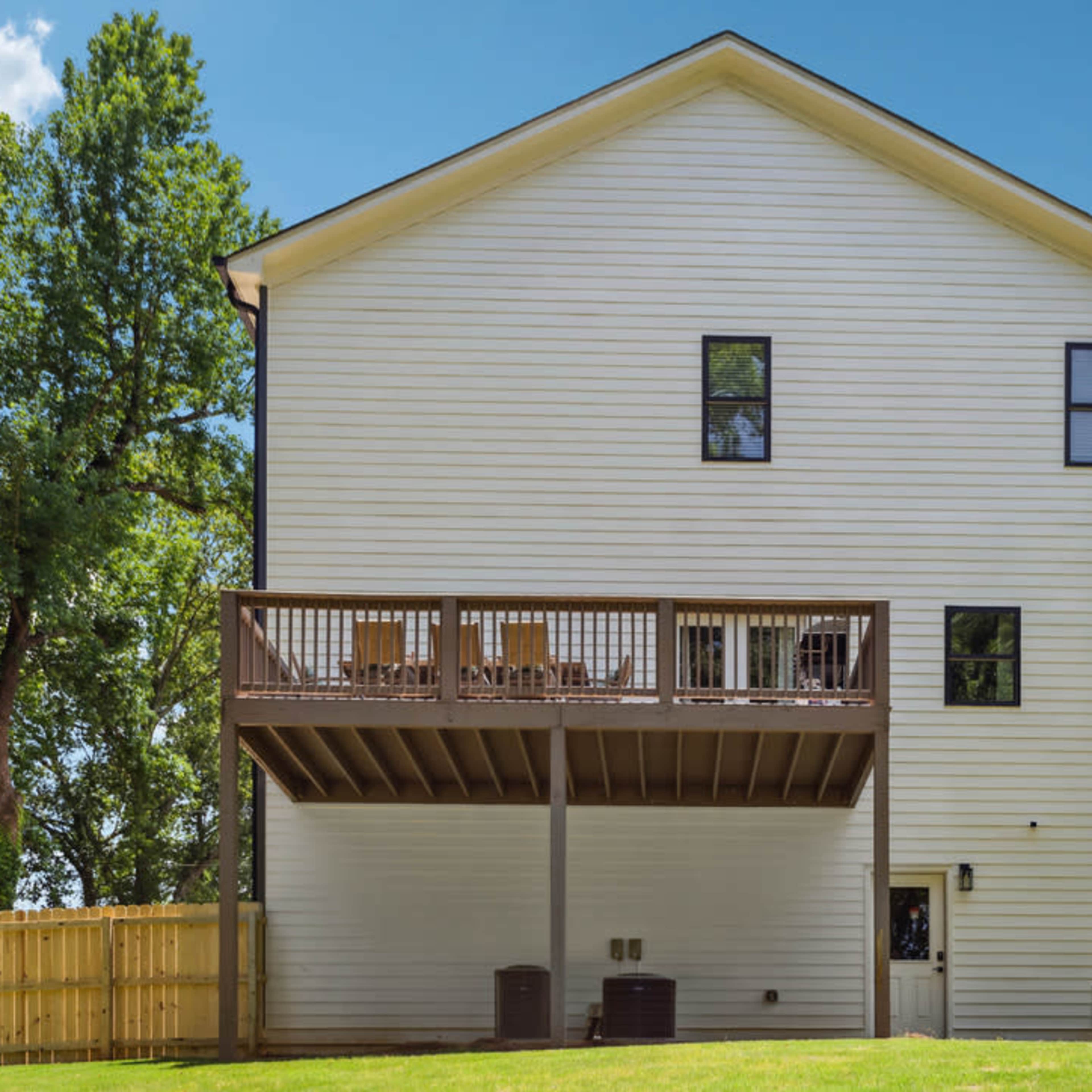 The back of a two-story house with a wooden deck and a fenced yard under a clear sky.