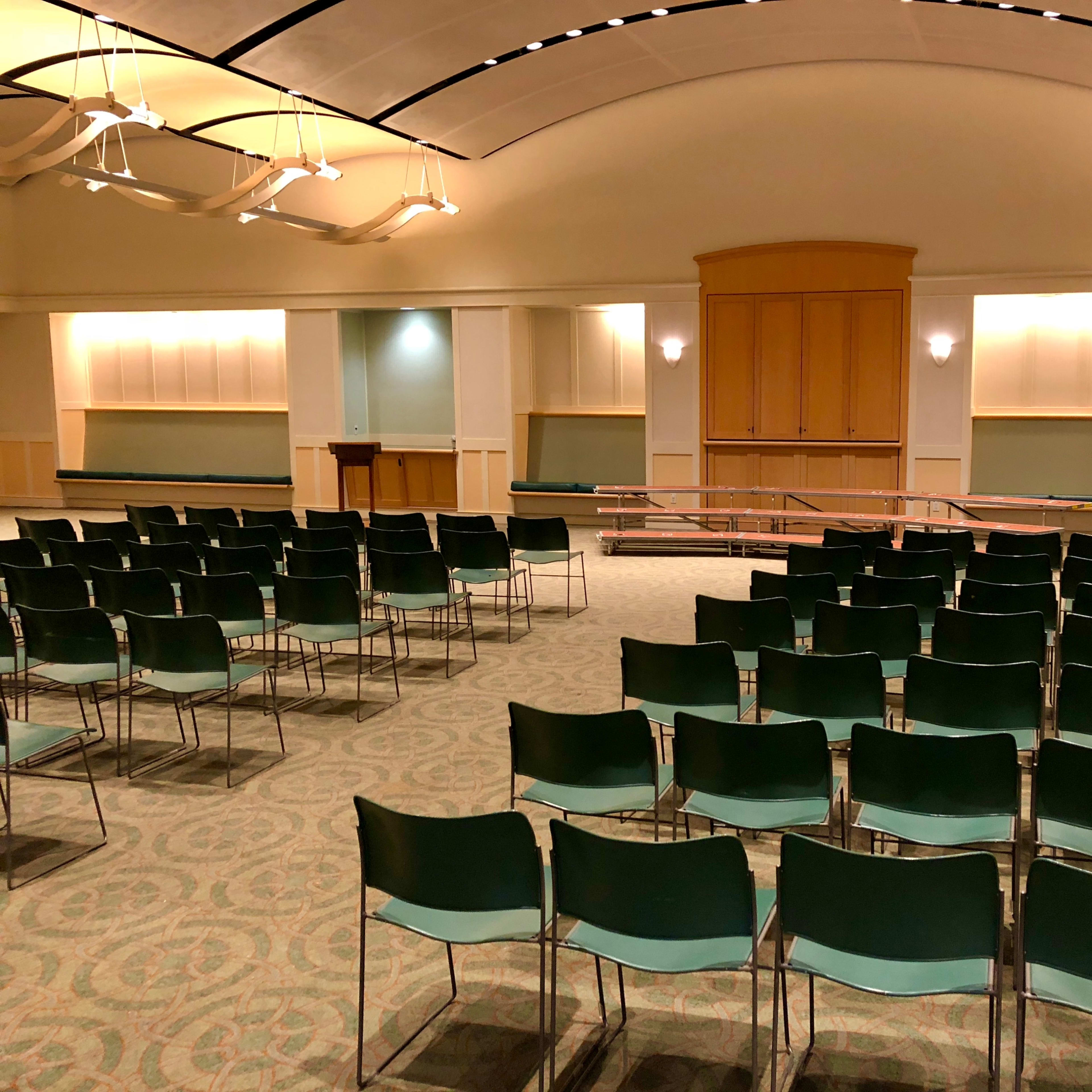 An empty indoor auditorium with rows of green chairs facing a wooden stage at the front.