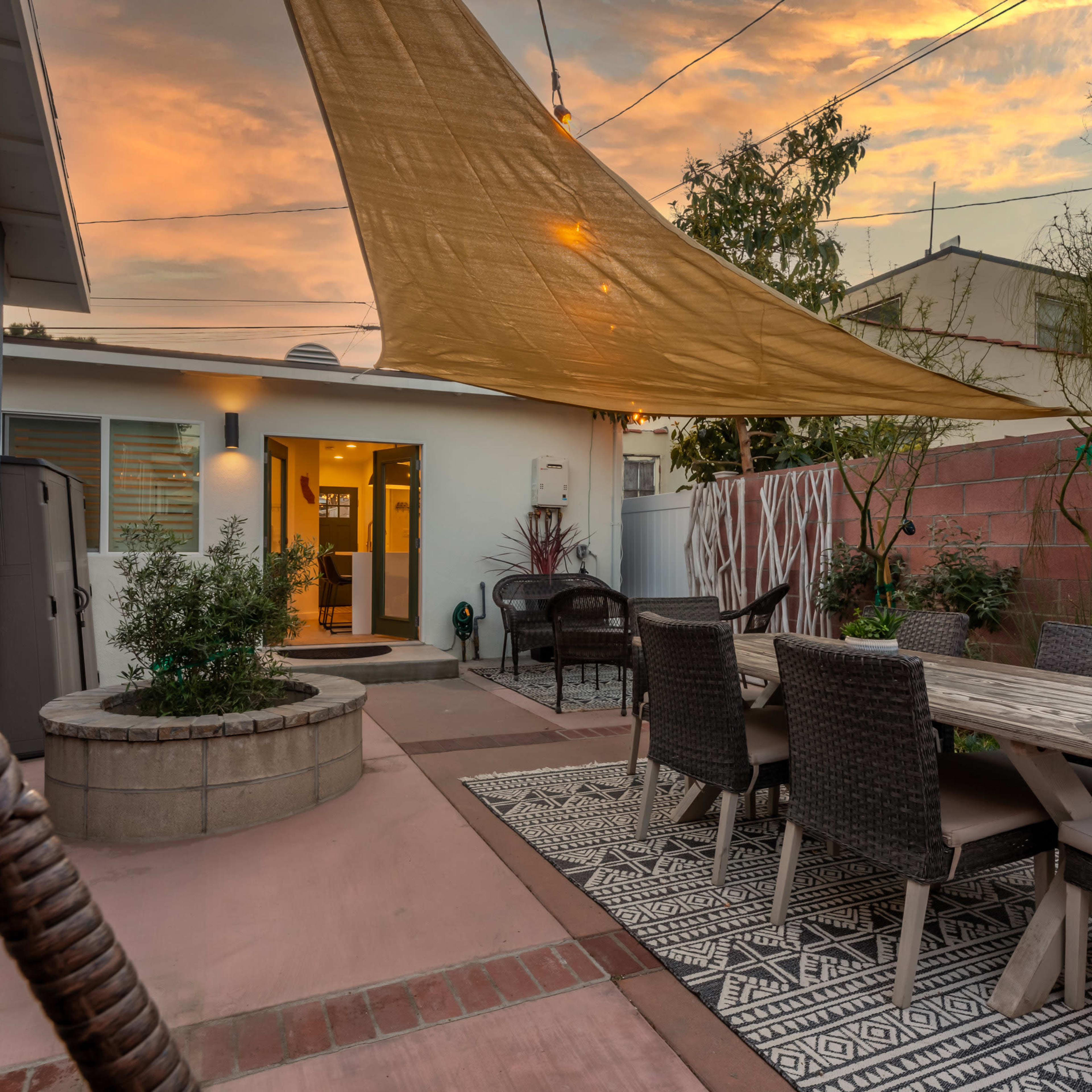 A backyard patio features a wooden dining table surrounded by wicker chairs, shaded by a large cloth canopy, with a small fire pit and decorative plants nearby under a colorful sunset sky.
