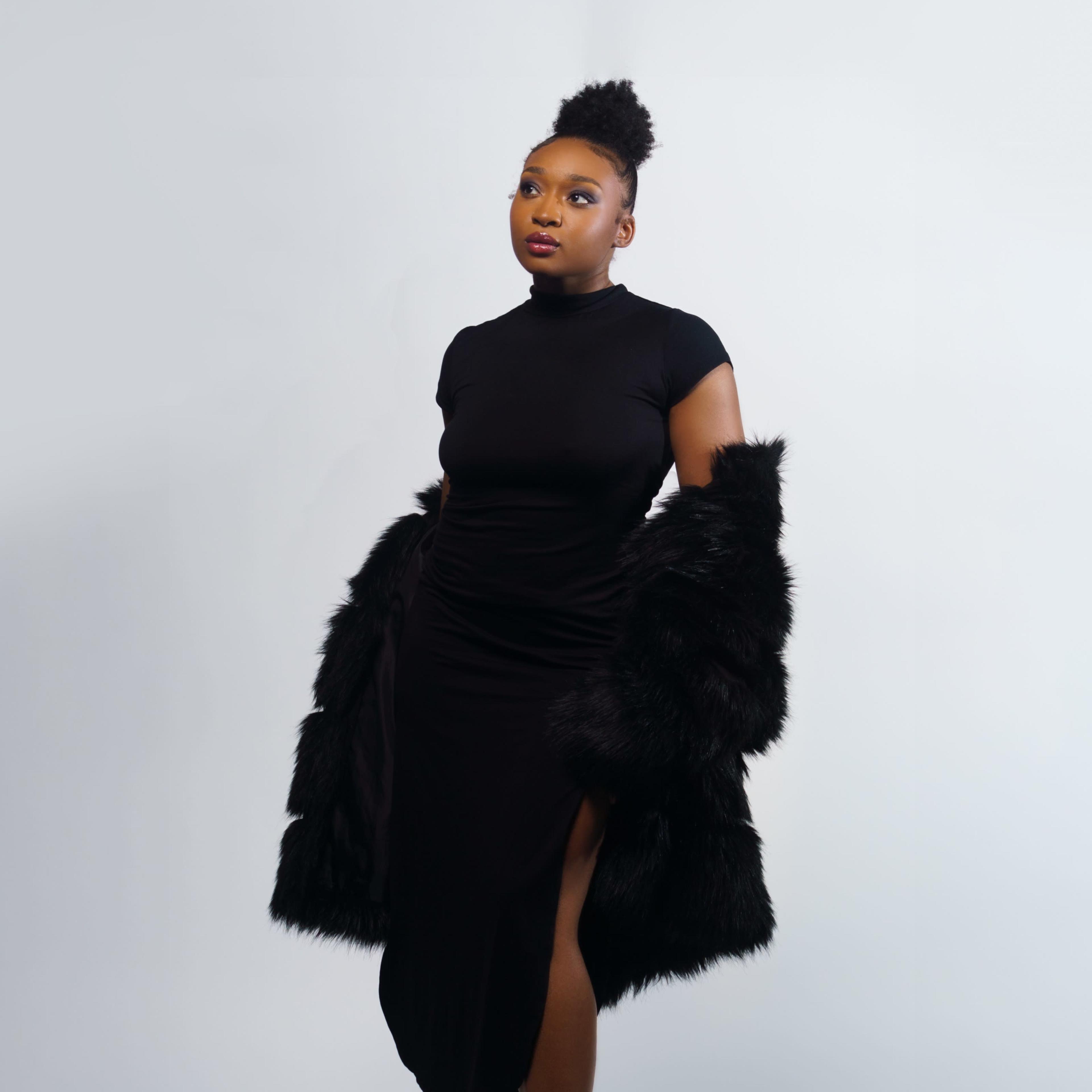 A woman poses in a black dress and fur coat against a plain background.