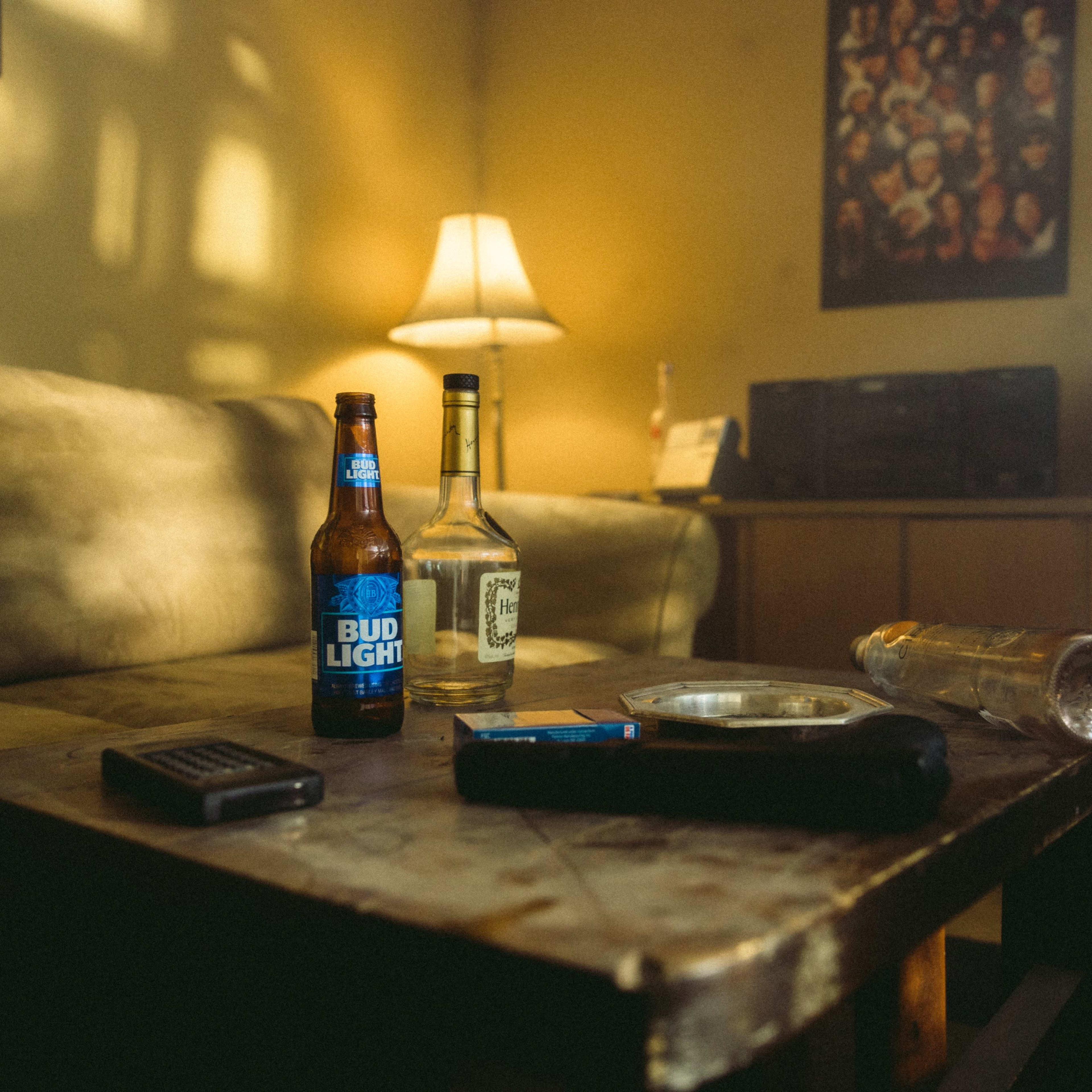 A small living room scene features a worn wooden coffee table with two bottles of beer, a liquor bottle, an empty glass, and a remote control, illuminated by a lamp in the background.