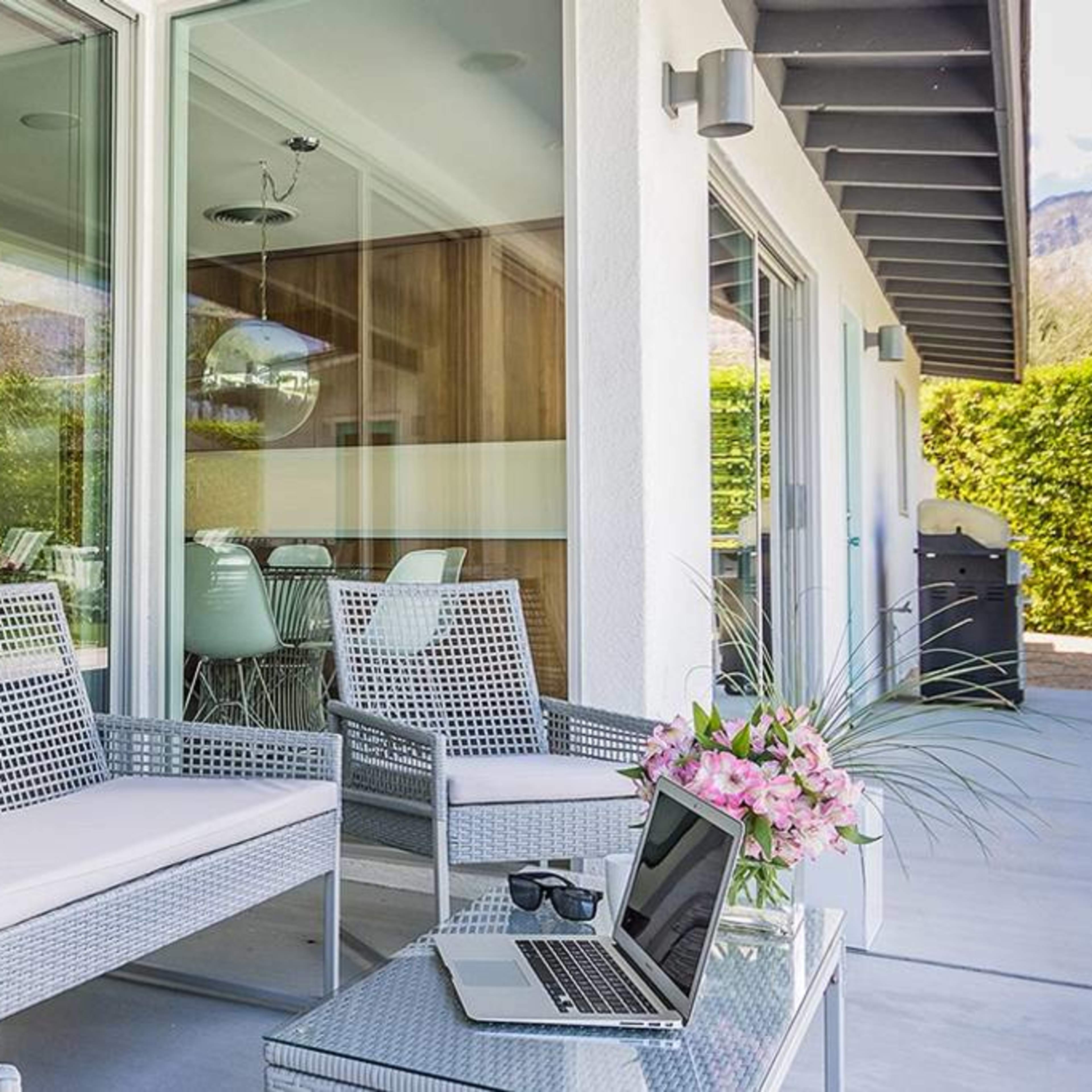 A modern patio features wicker furniture, a laptop on a small table, and a flower arrangement, with a view of a green hedge and glass doors.