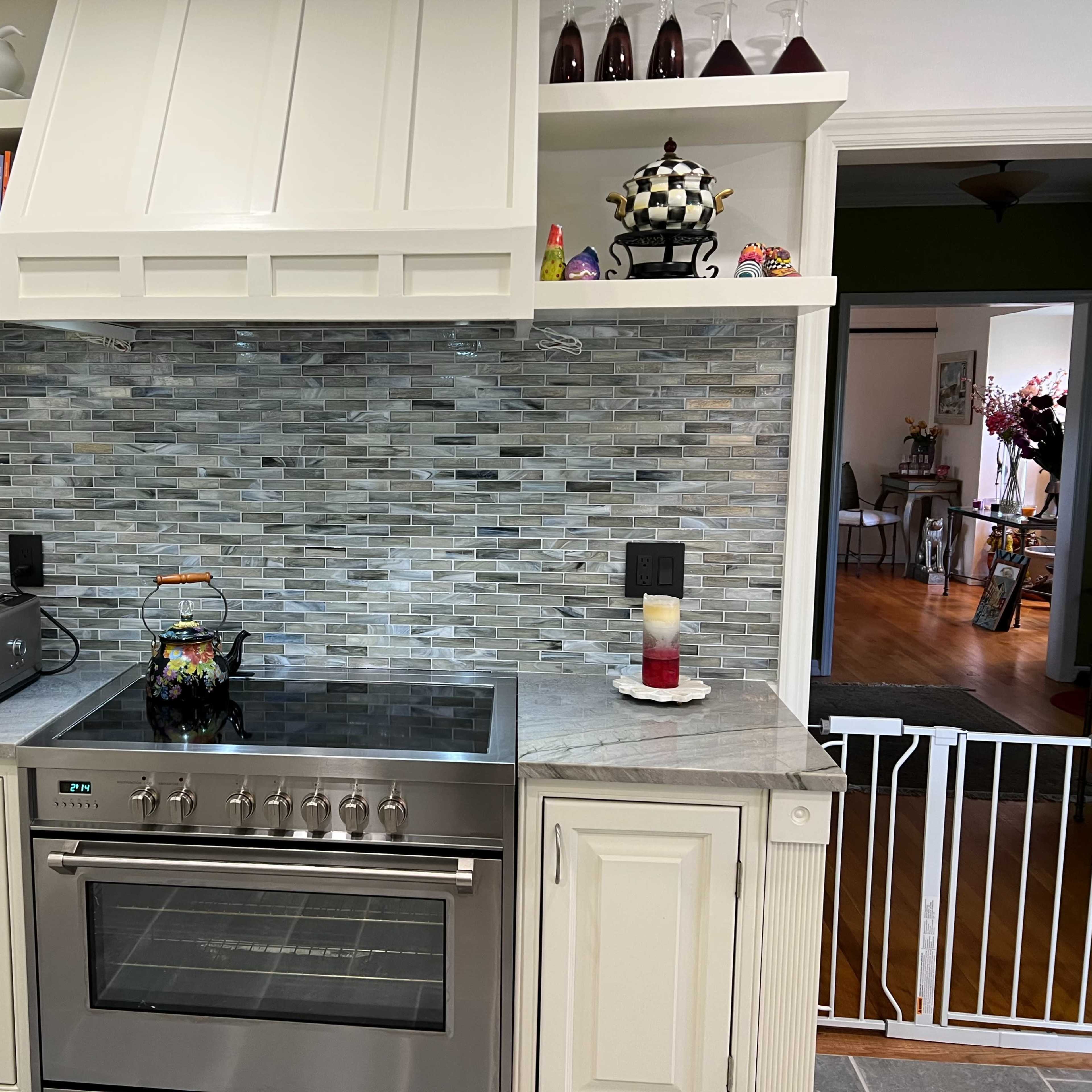 A kitchen with a stainless steel stove, a gray stone backsplash, and white cabinetry featuring open shelves with decorative items.