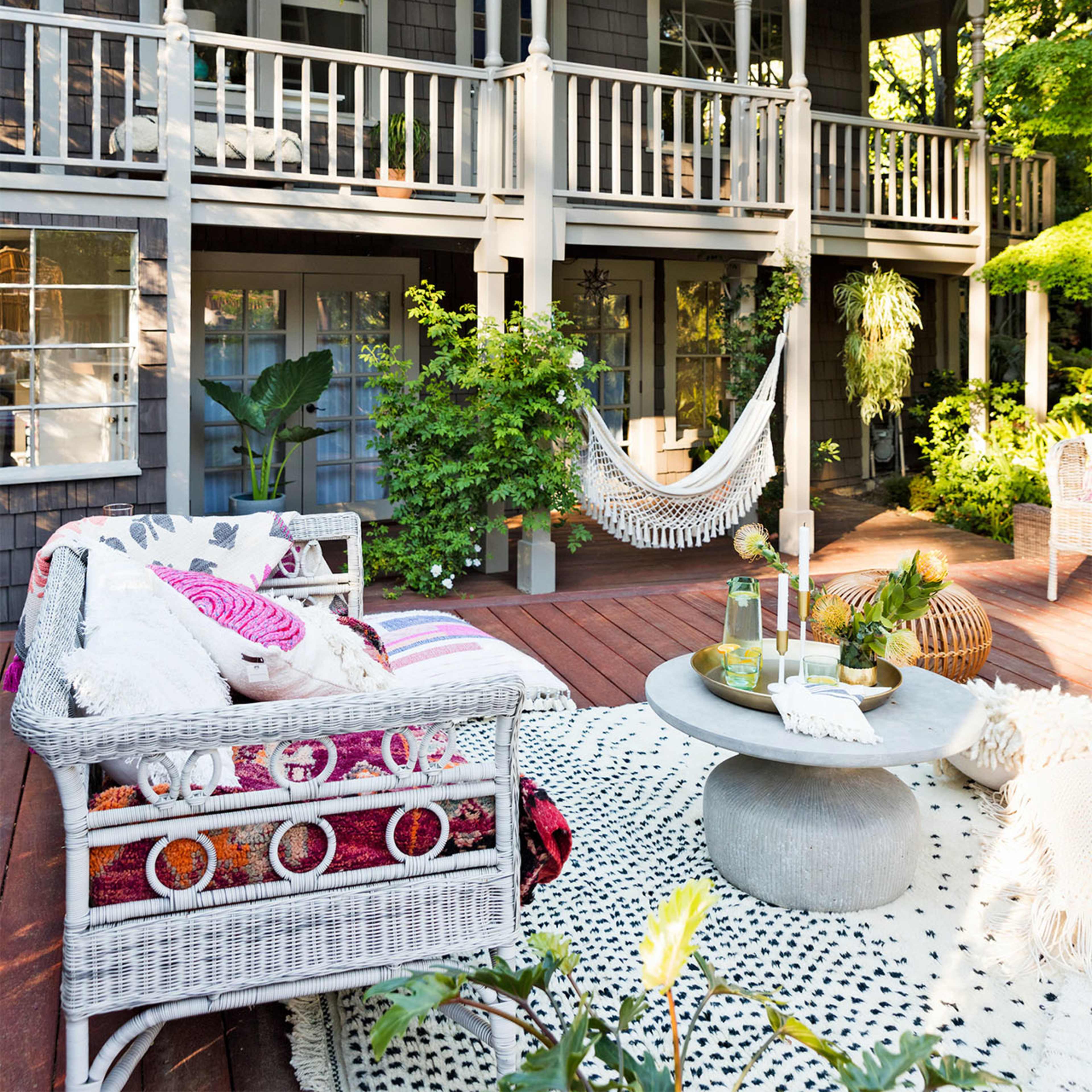 The scene features a cozy outdoor patio with a hammock, comfortable seating, and a central table, surrounded by greenery and a two-story house in the background.