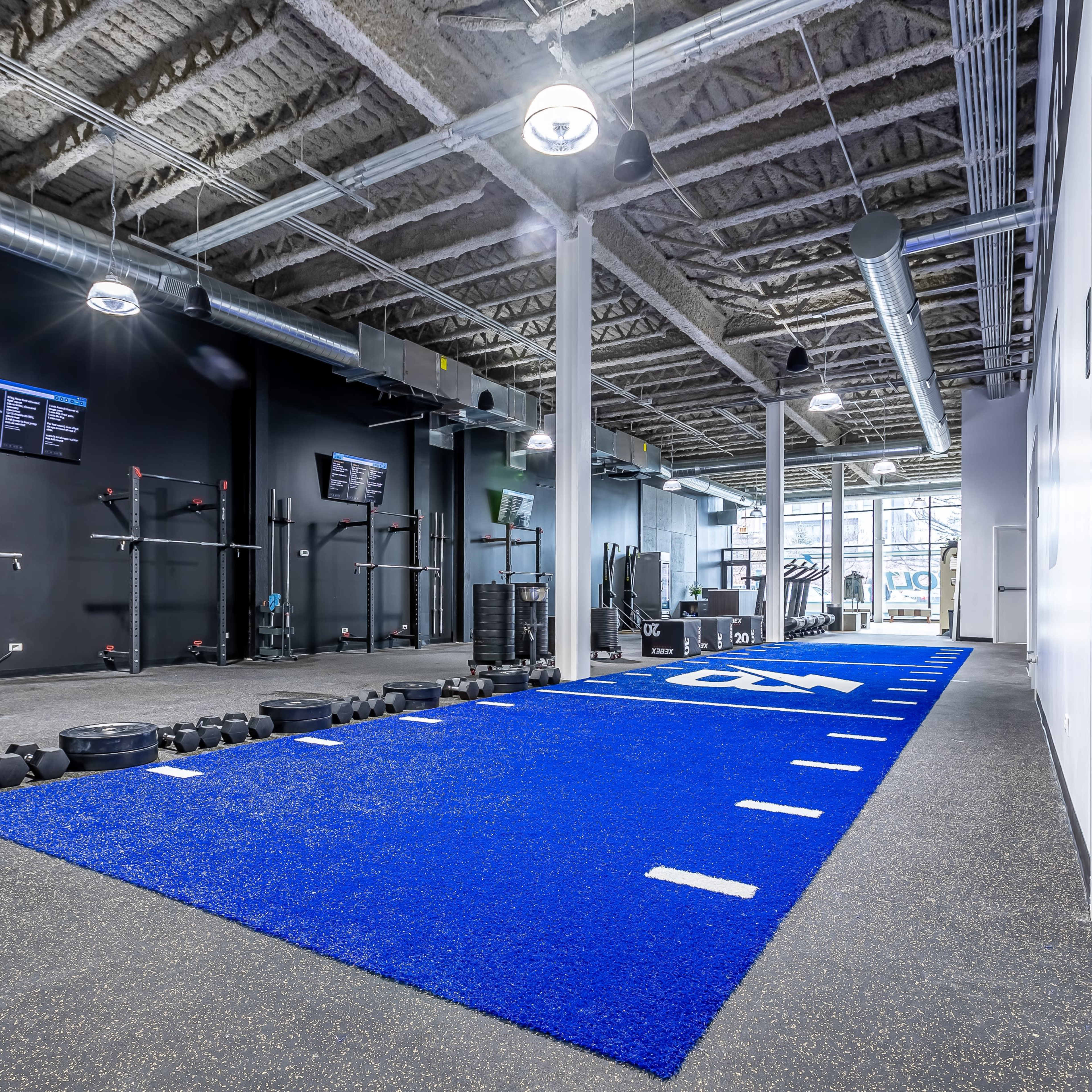 A modern gym interior featuring a long blue turf area with fitness equipment and weights along the sides.