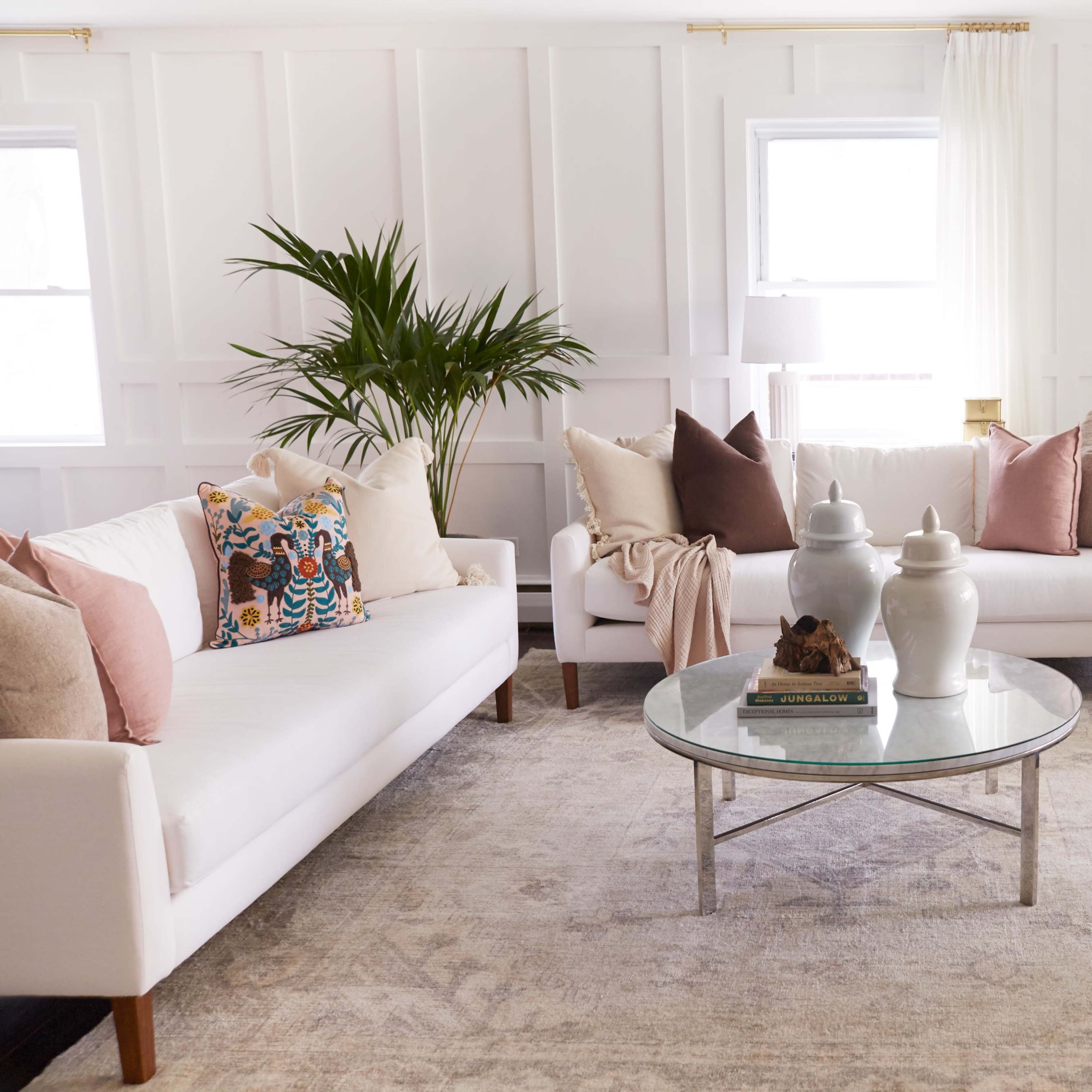 A bright, stylish living room features two white sofas with decorative pillows, a glass coffee table, and a soft area rug, surrounded by plants and natural light.
