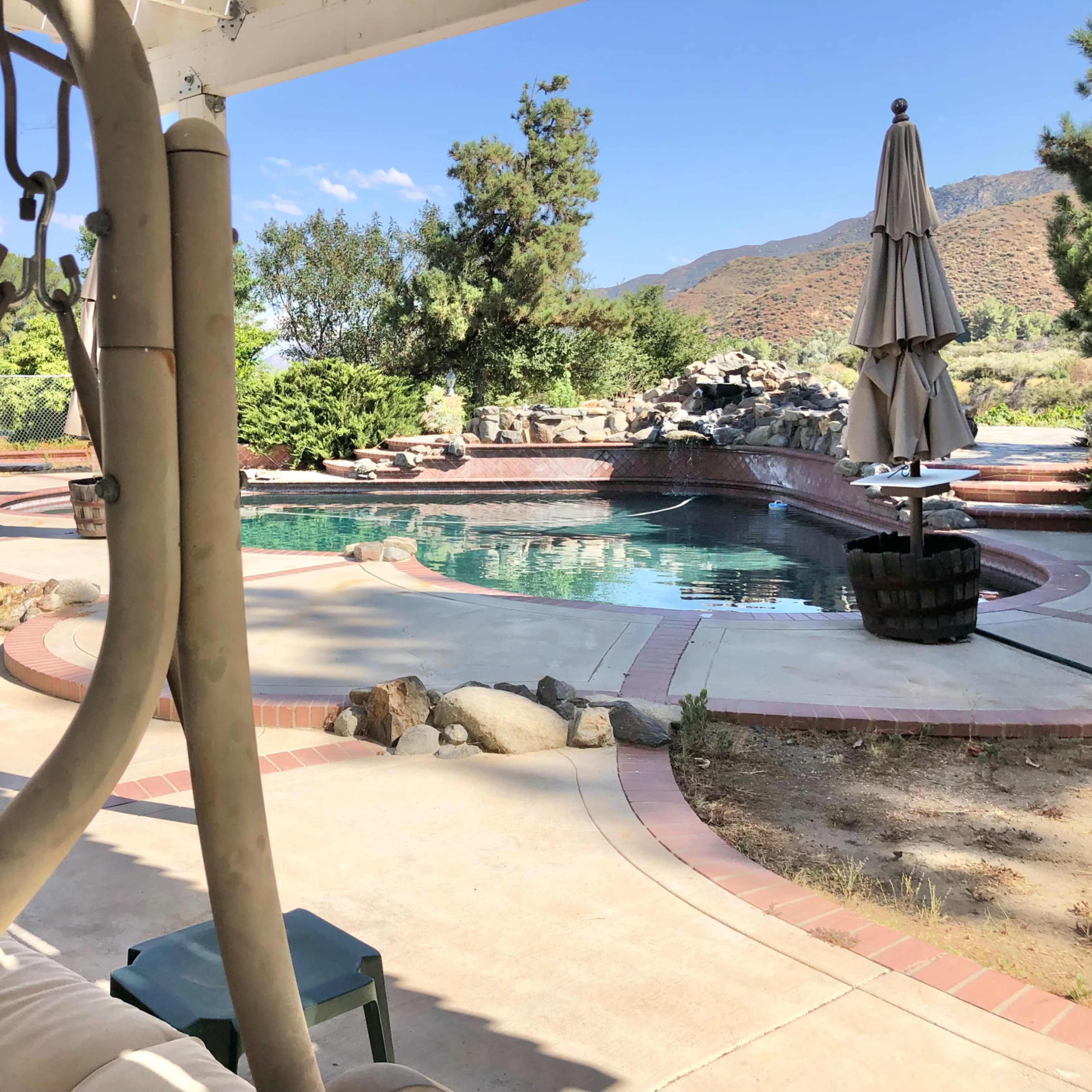 A backyard with a swimming pool surrounded by landscaping, a patio area with a swing, and a large umbrella near the water's edge.