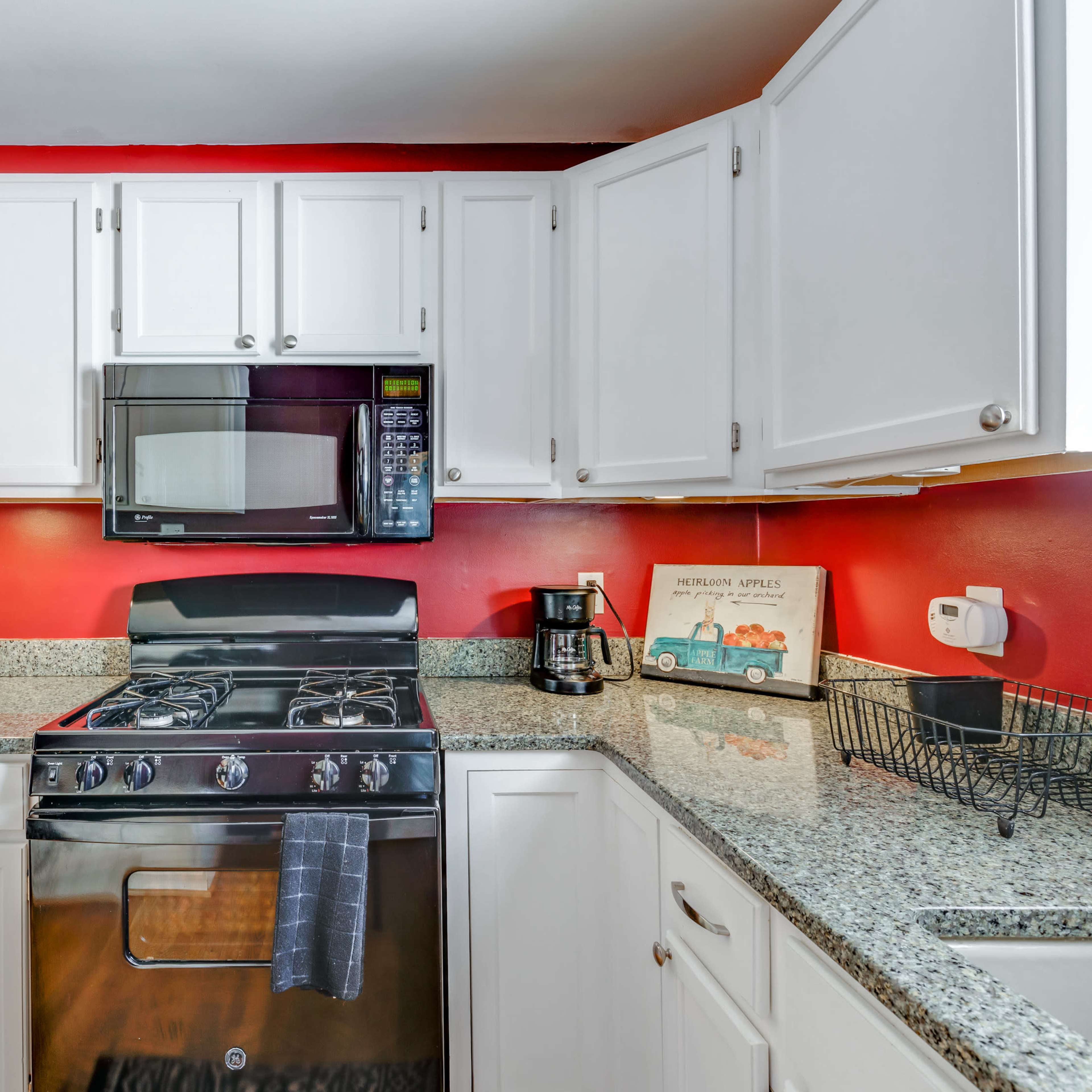 The kitchen features white cabinets, a black stove and microwave, a coffee maker, and a granite countertop with a red wall backdrop.