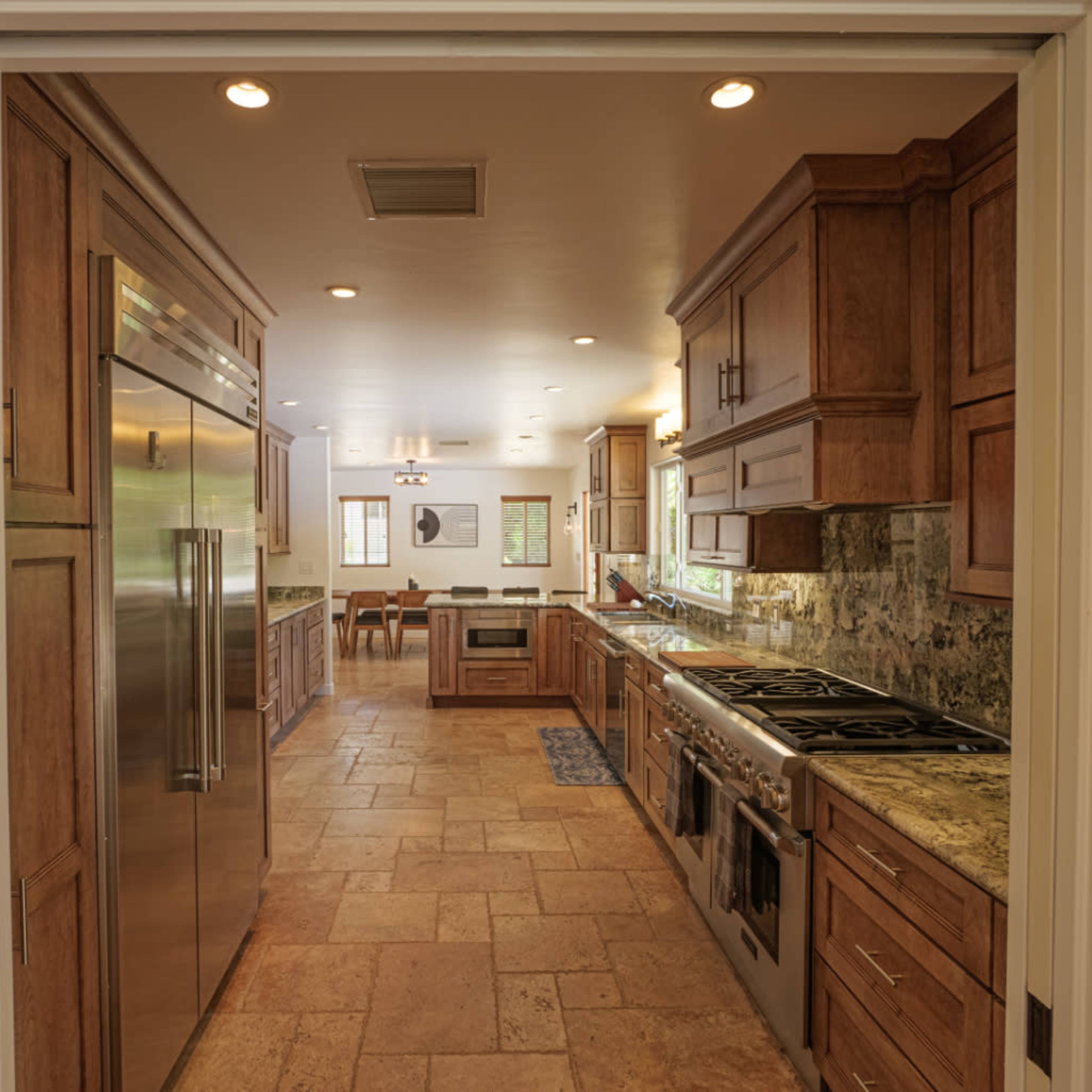 A spacious kitchen with wooden cabinets, stainless steel appliances, and a tiled floor, visible through an open doorway.